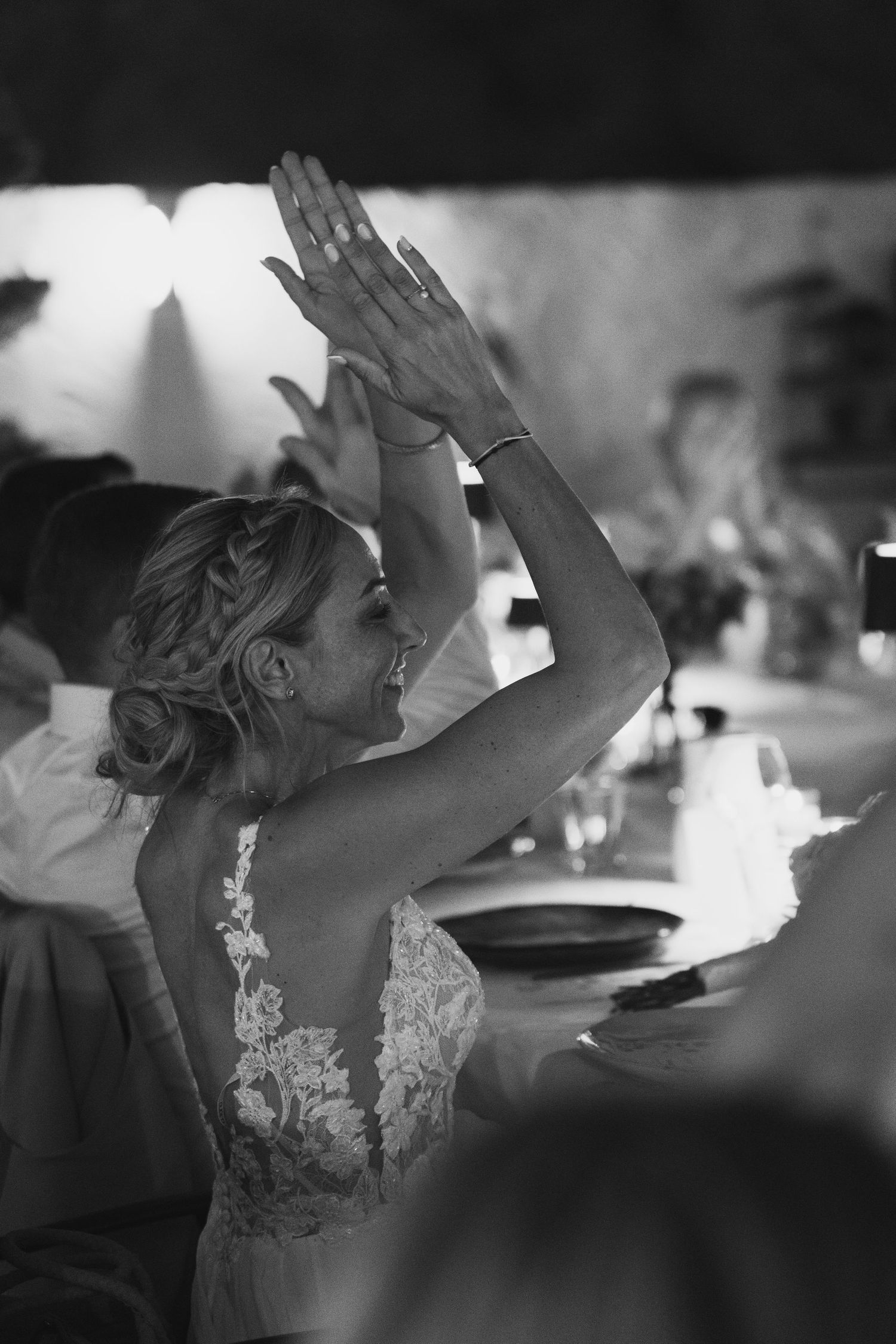 A person in a white lace dress raises their hands in celebration during what appears to be a wedding reception.