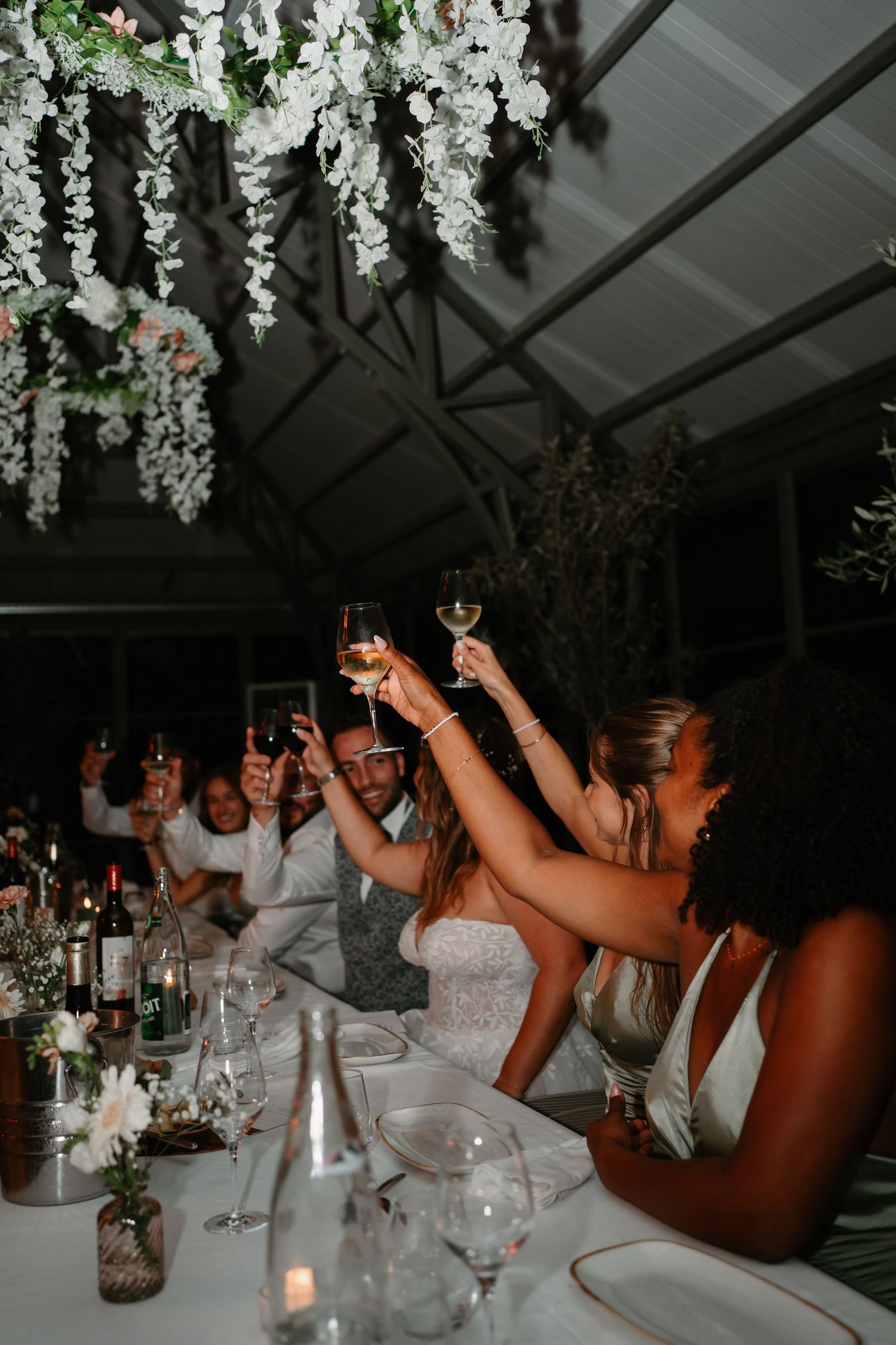 Wedding reception guests raise their glasses in a toast under hanging white floral decorations at an elegant dinner party.