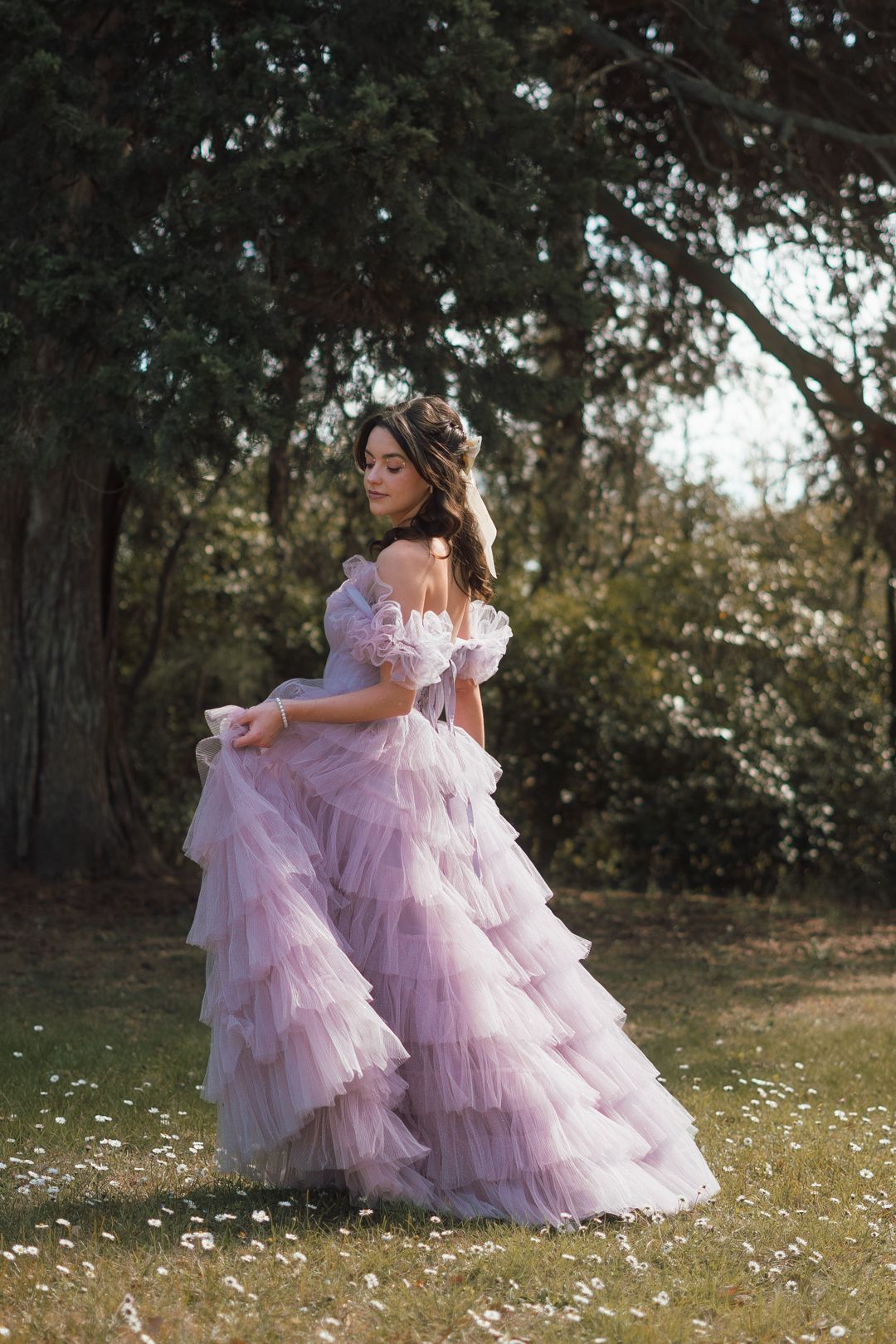Two figures in matching pink tulle gowns embrace in a sunlit meadow with white wildflowers and trees in the background.