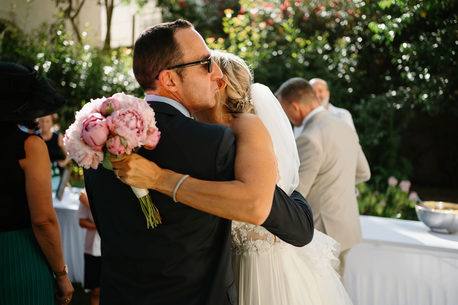A tender embrace between newlyweds at an outdoor garden wedding ceremony with pink floral bouquet details.