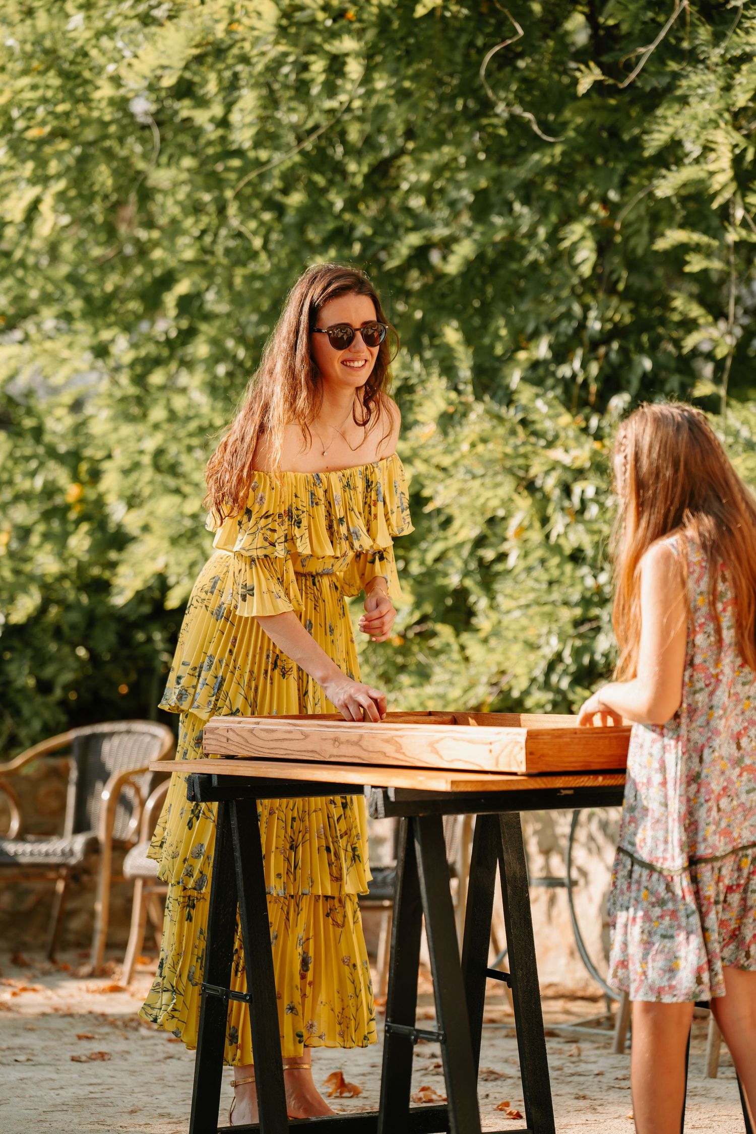 Two people playing backgammon at an outdoor table surrounded by lush green trees on a sunny day.
