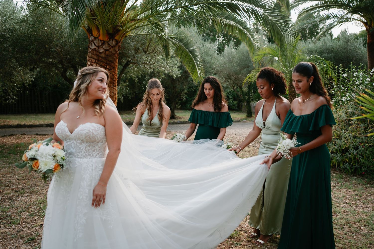 Bridesmaids in emerald green dresses help adjust a bride's flowing white dress in a tropical garden setting.