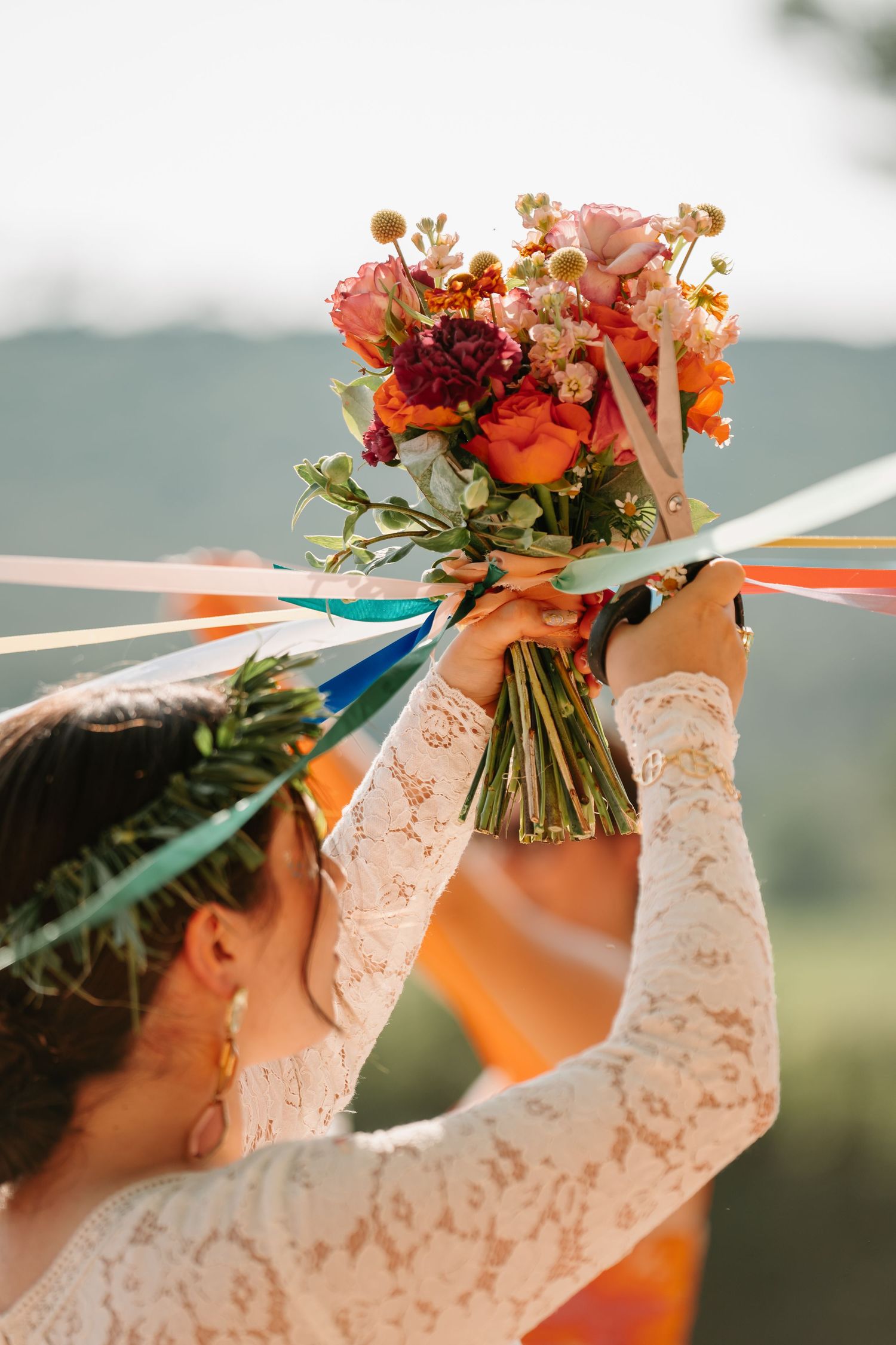 A person in white lace sleeves holds up a vibrant orange and red floral bouquet against a blurred outdoor background.
