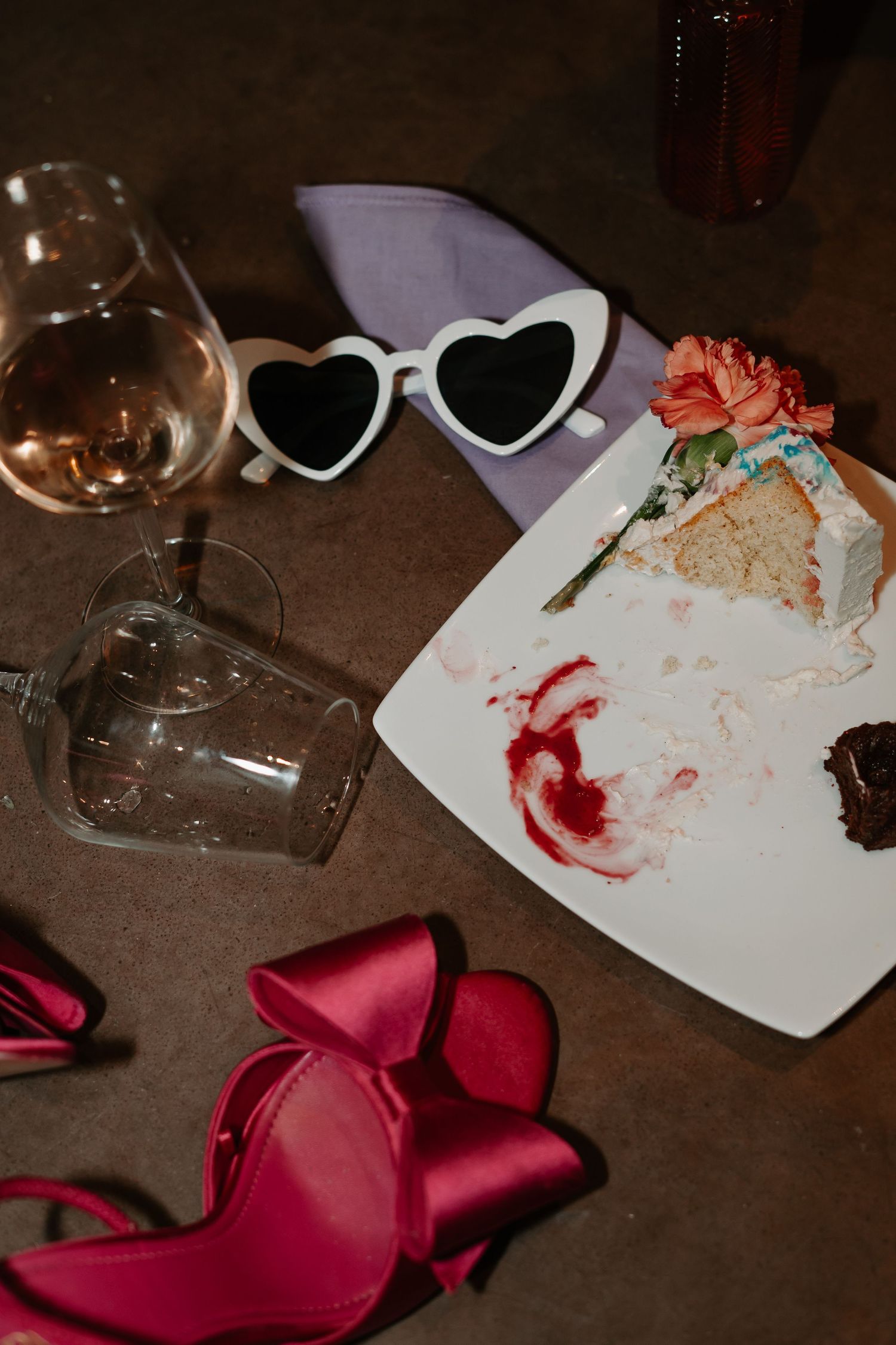 Heart-shaped sunglasses, wine glass, red high heels, and a plate with cake crumbs create a romantic party scene.