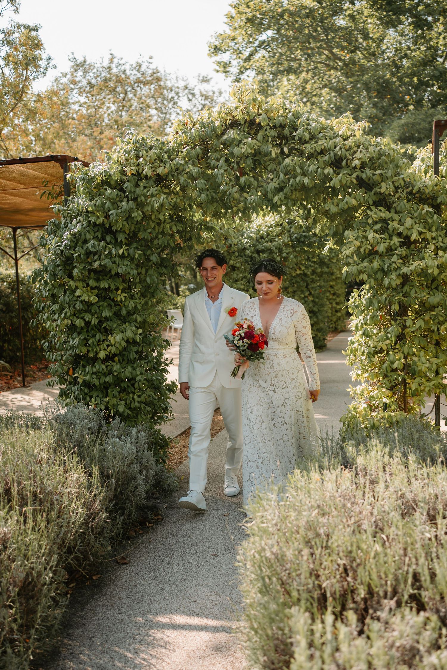 A couple walks down a garden path surrounded by lush greenery and flowering plants on a sunny day.