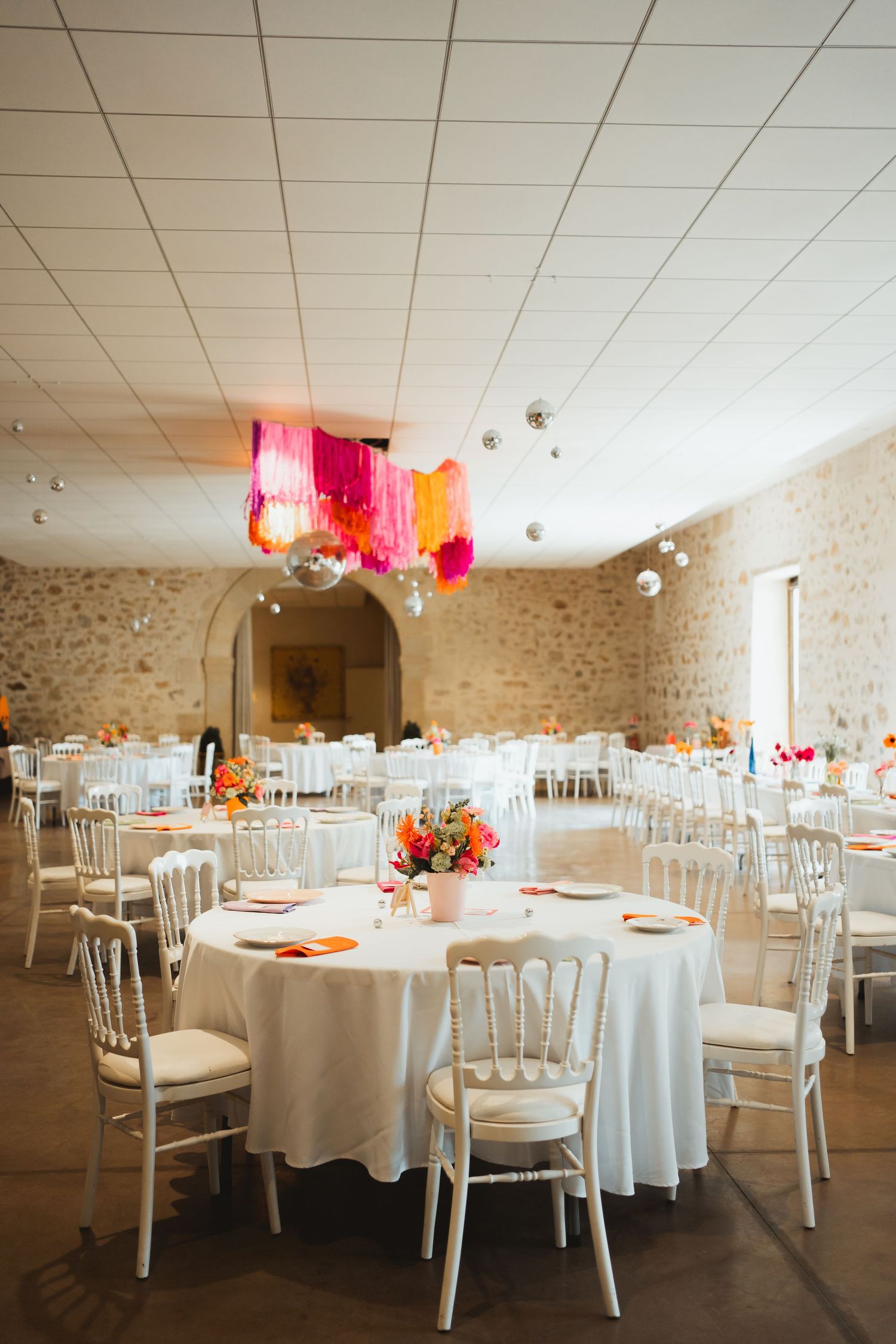 Elegant wedding reception venue with white tables, chairs and pink floral centerpieces in a rustic stone-walled room.