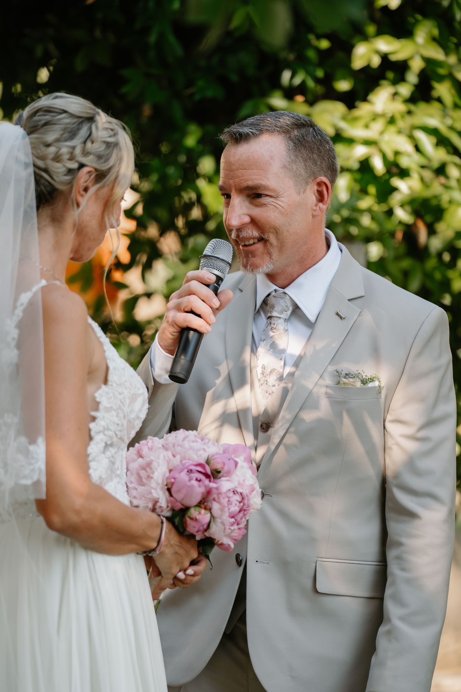 A tender first look moment between bride and groom in light gray suits with pink peony bouquet in outdoor setting.