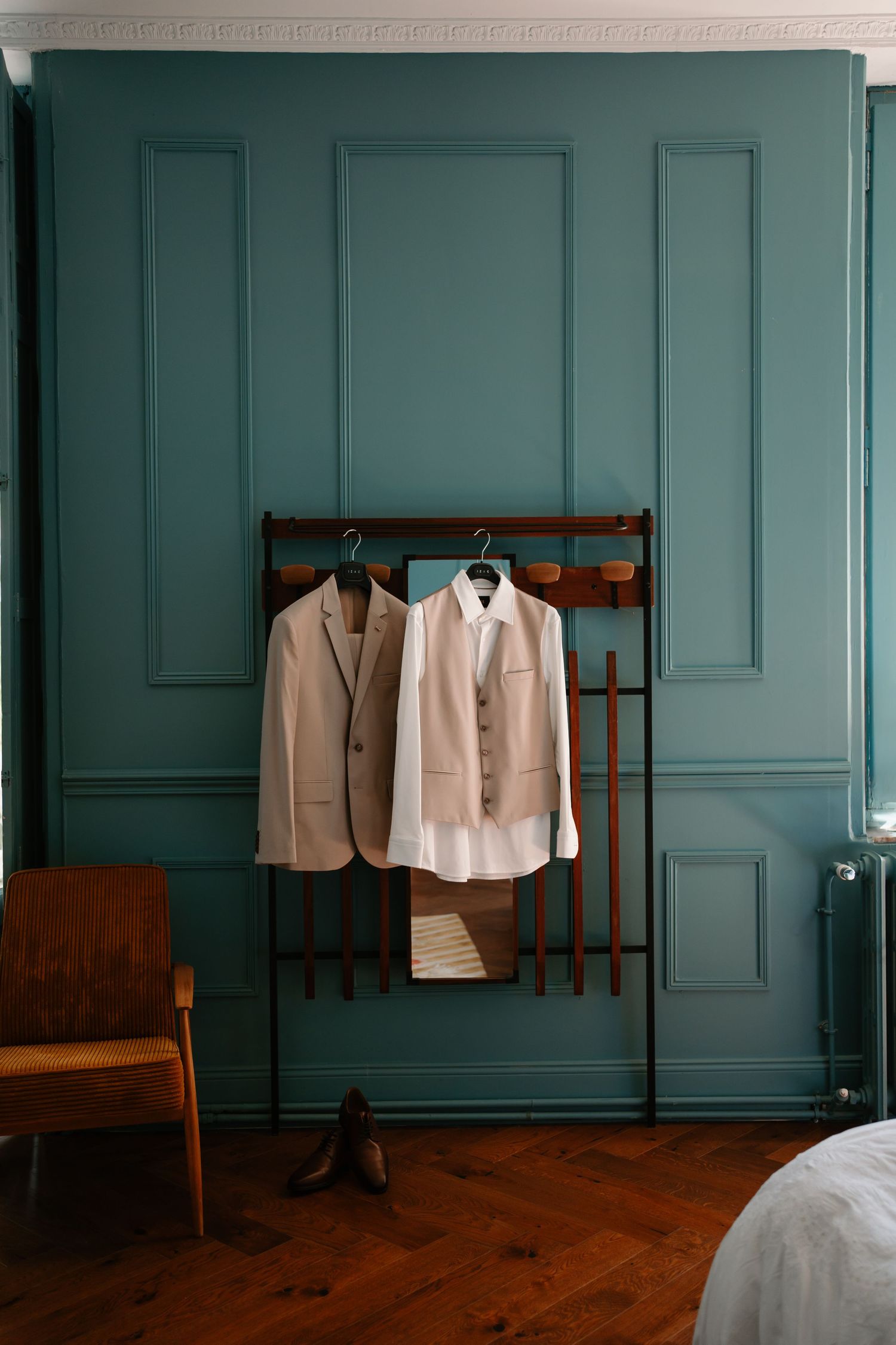 White dress shirt and beige suit jacket hanging on wooden coat rack against teal paneled wall in minimalist room.