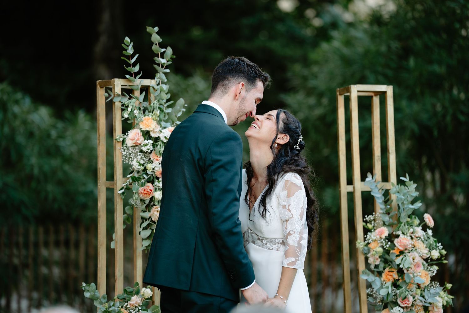 Romantic couple embraces during outdoor garden wedding ceremony with wooden arch and floral decorations.