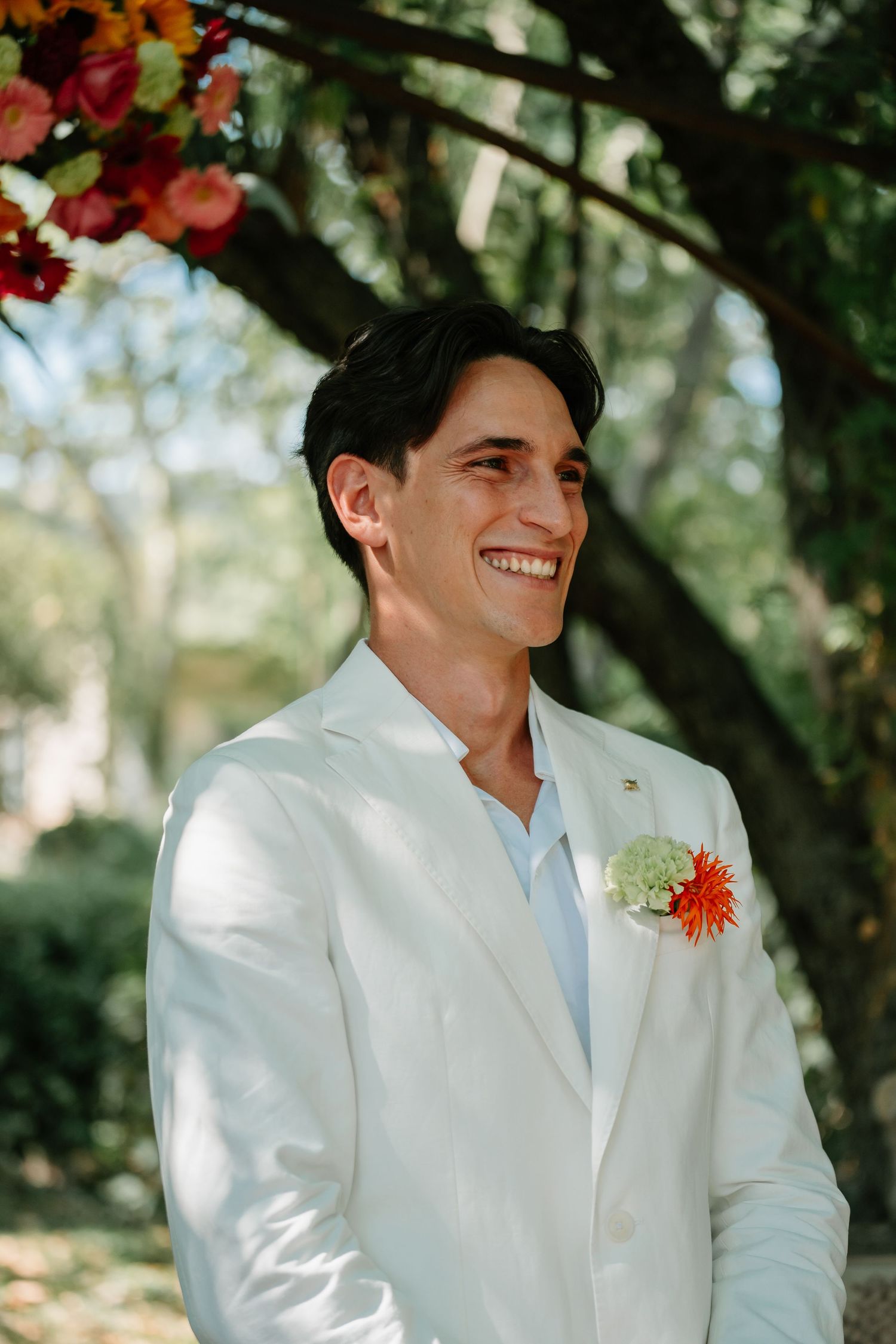 Groom in a crisp white suit with light blue tie and orange boutonniere smiles during outdoor wedding ceremony.
