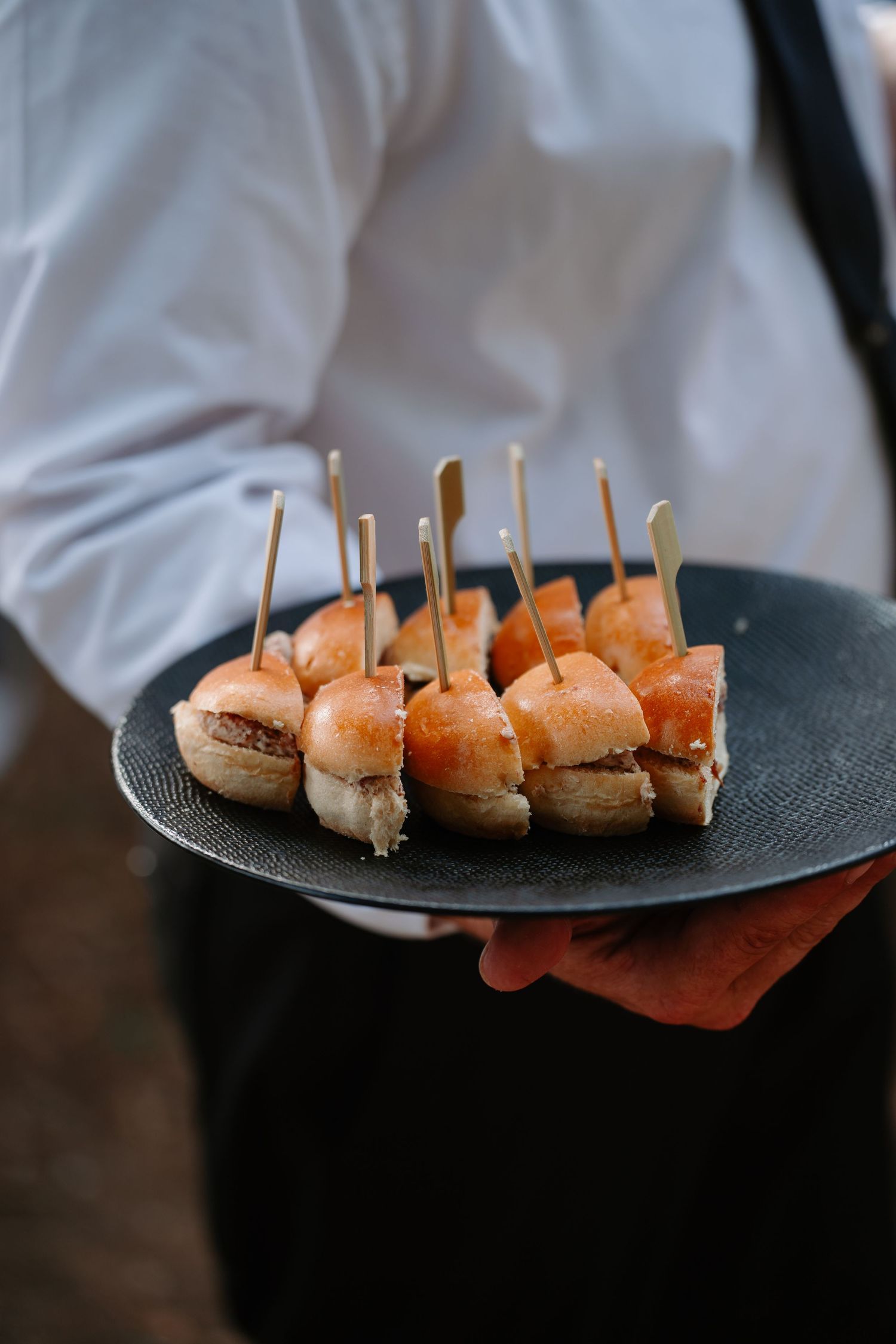 A server holds a black plate with mini burger sliders topped with wooden toothpicks at a catered event.