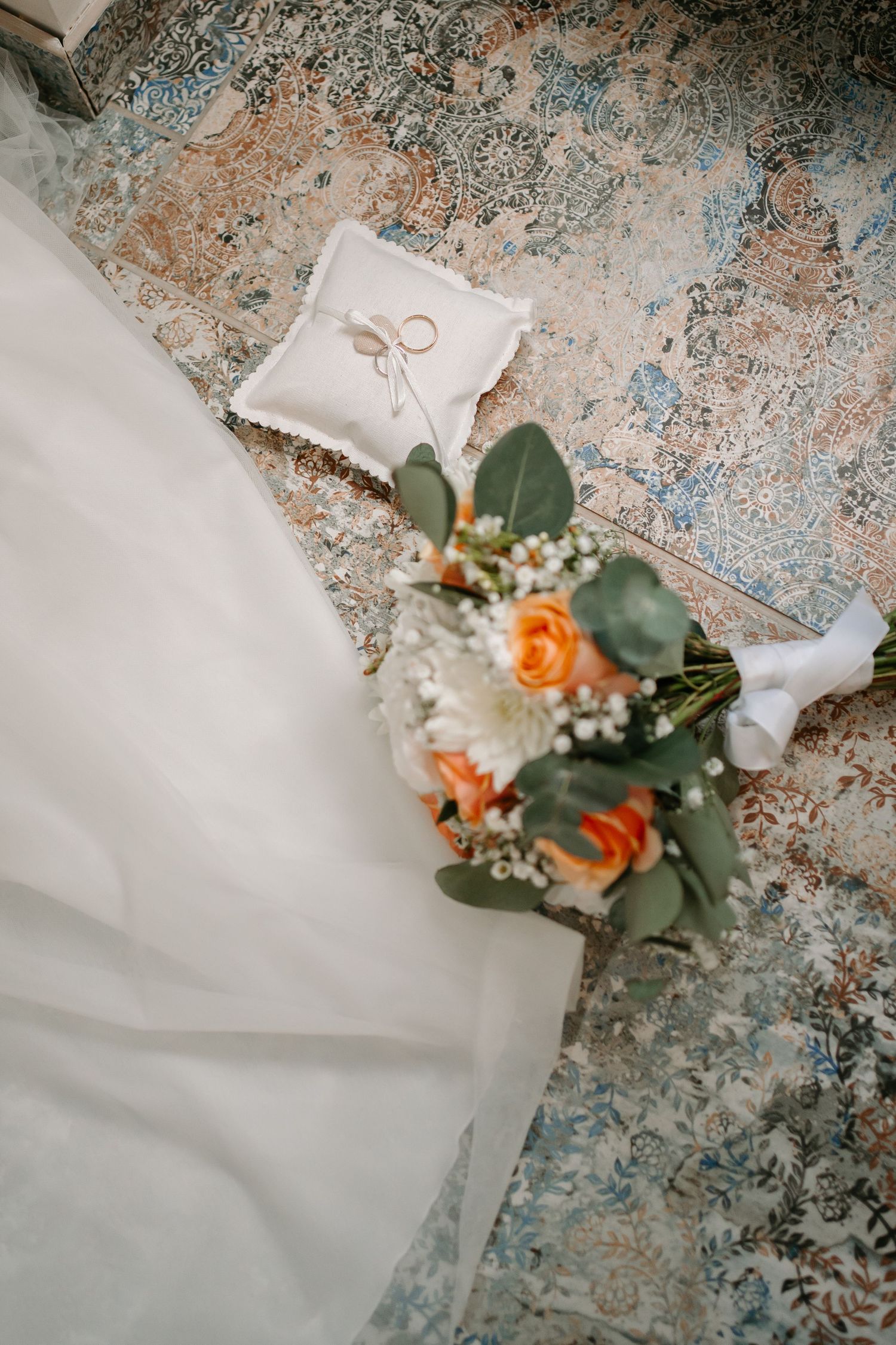 A delicate orange rose with greenery and baby's breath rests on flowing white fabric against a distressed blue surface.