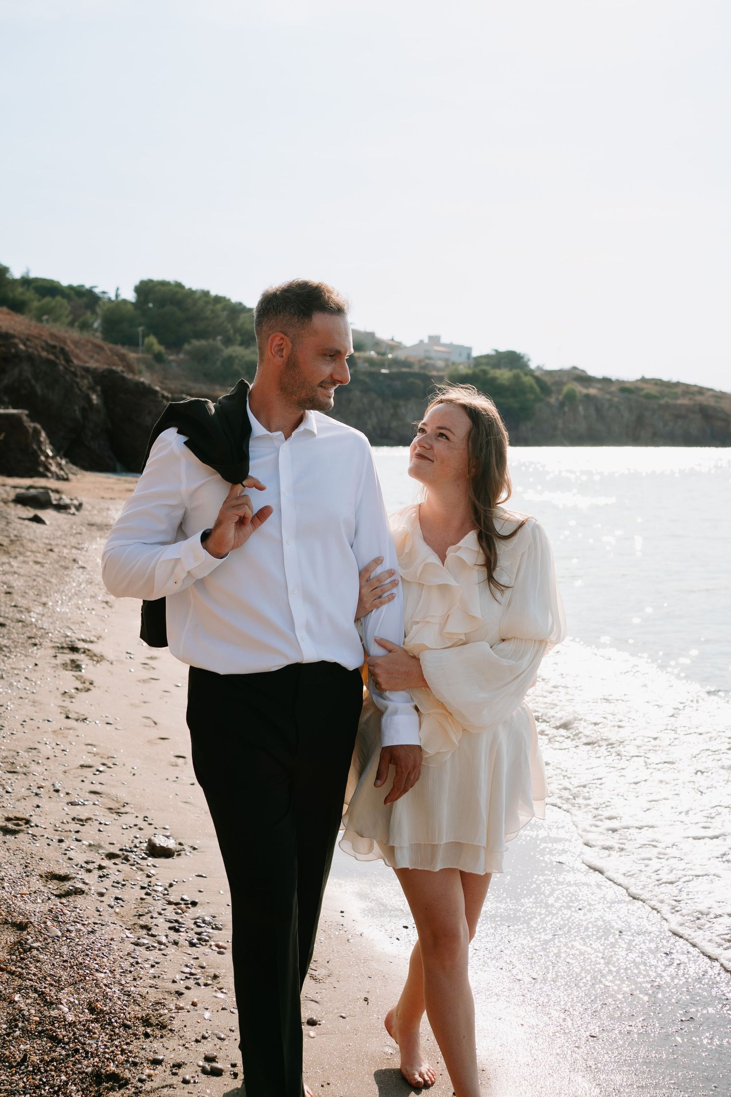 A happy couple walks hand-in-hand along a sunny beach, man in white shirt holding jacket, woman in white dress smiling.