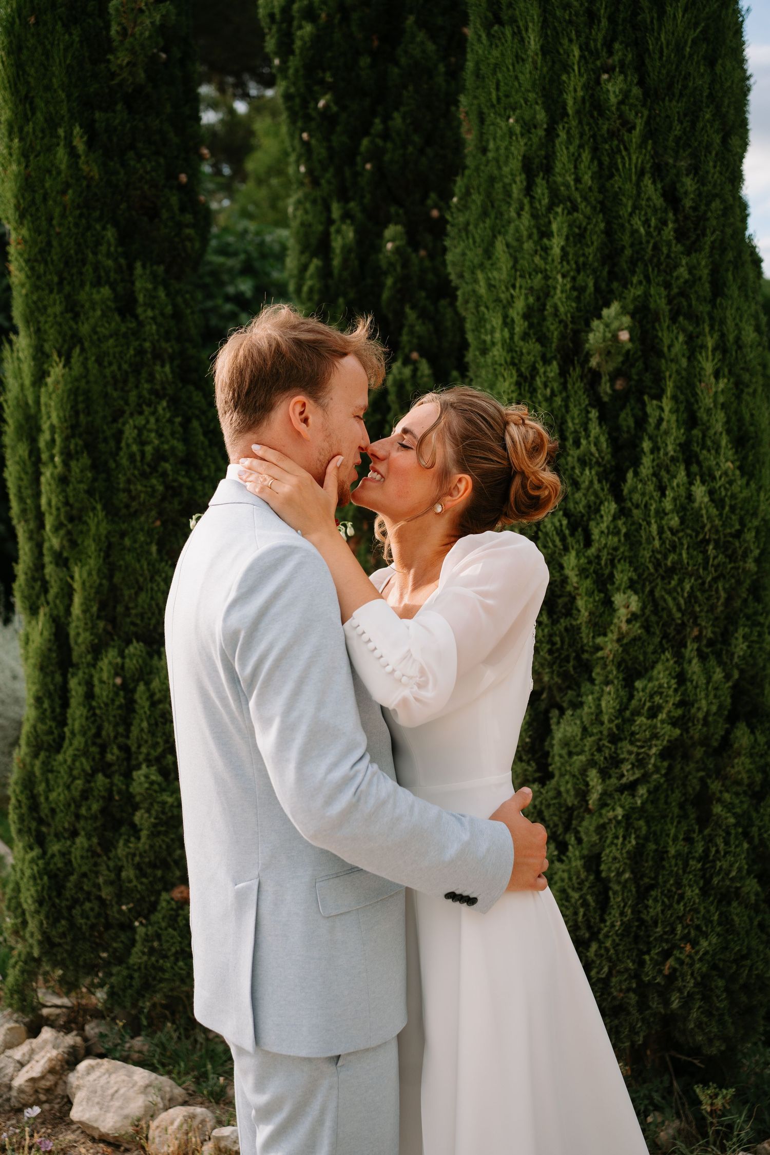 A bride and groom share a romantic moment outdoors, embracing near tall cypress trees in wedding attire.