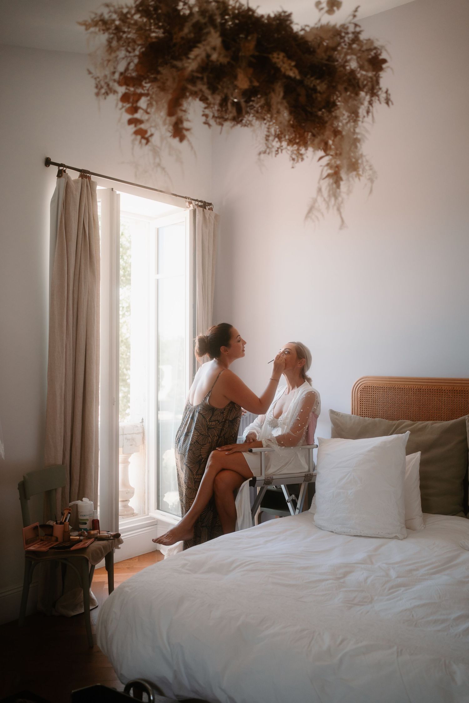 A dreamy bedroom scene with soft natural light streaming through curtains, featuring a bed and dried pampas grass ceiling decor.