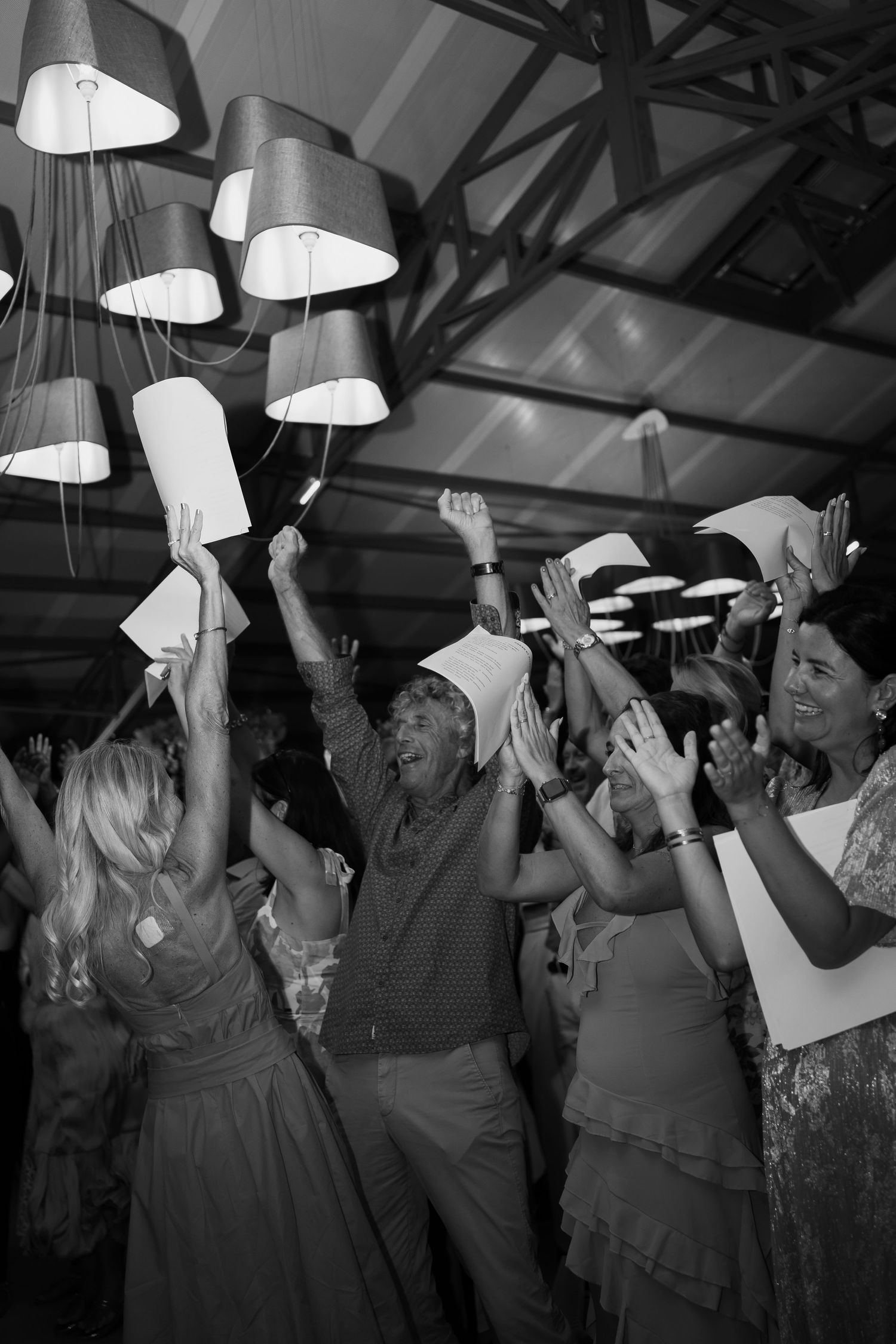 Wedding party guests dance and celebrate under hanging pendant lights at an indoor reception venue.