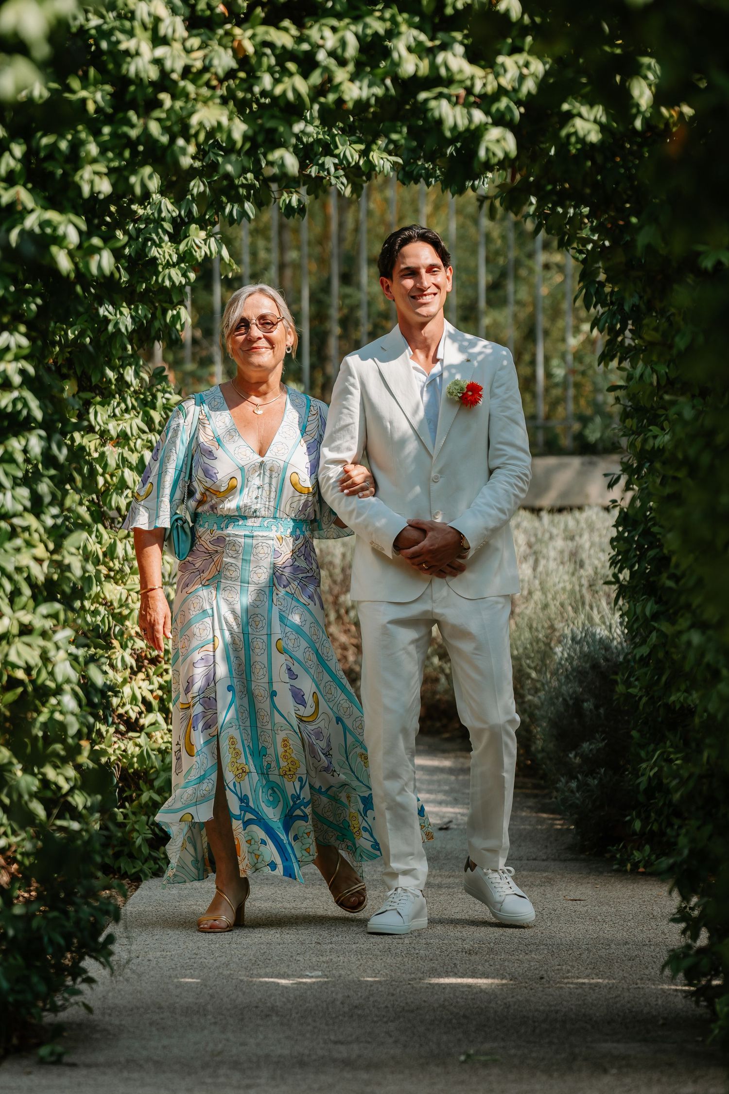 A couple shares a joyful moment walking together through a garden pathway in wedding attire.
