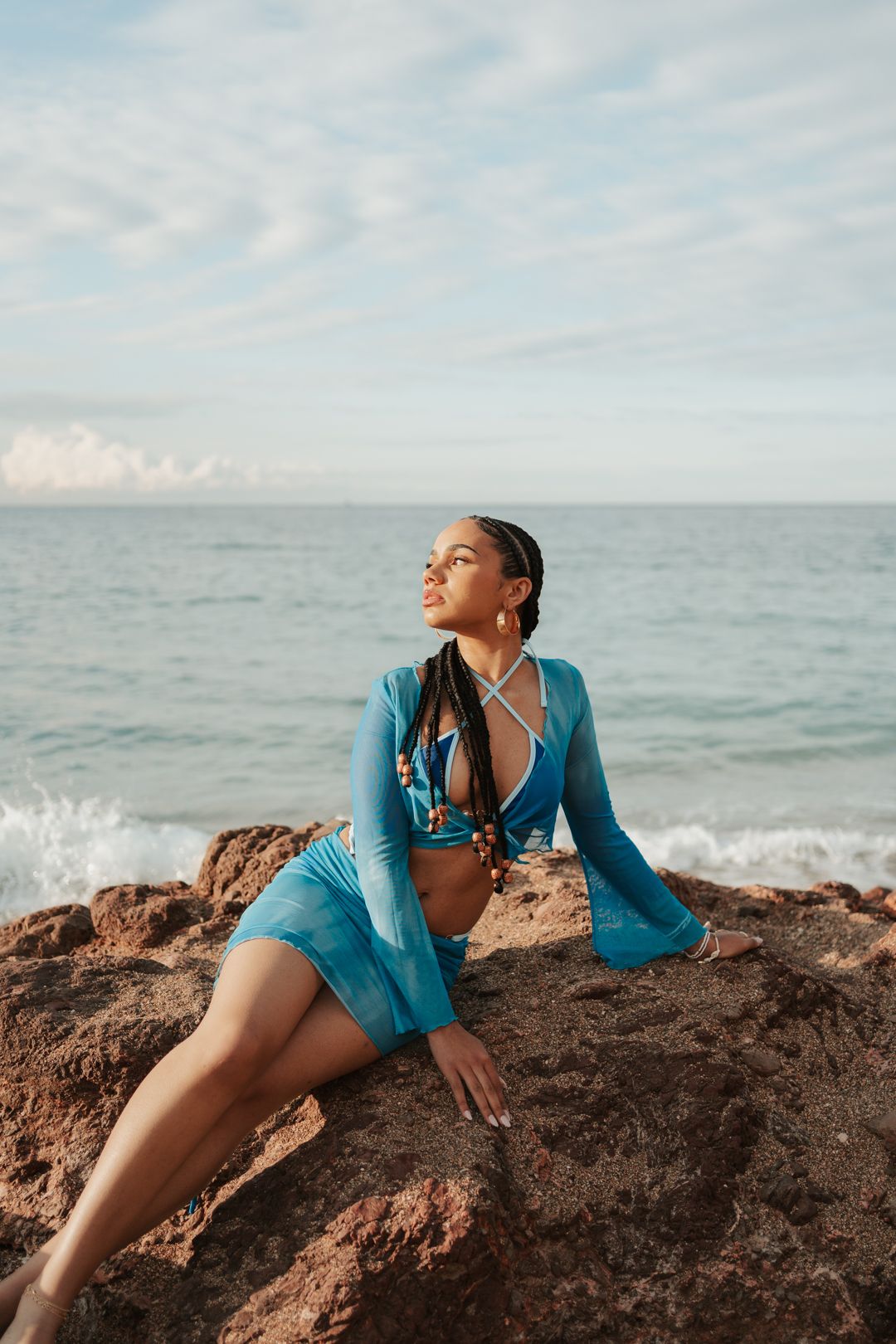 A figure in turquoise lounges on coastal rocks by the ocean at sunset.