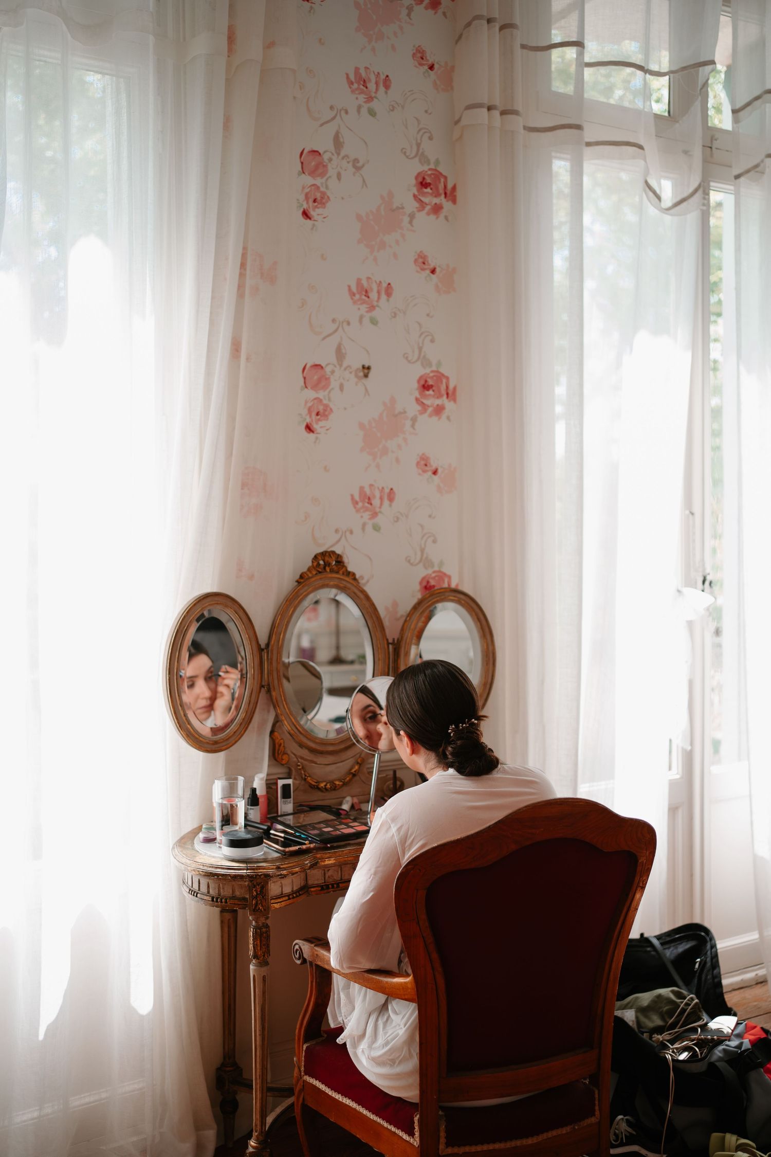 Person sitting at vintage vanity mirror with floral wallpaper in soft natural light.