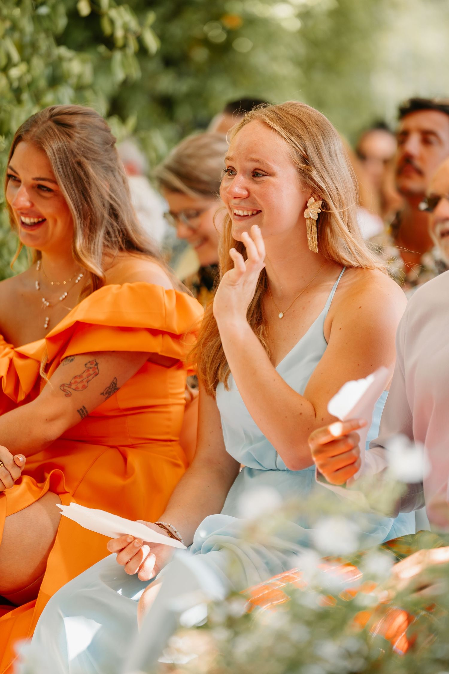 Wedding guests in orange and light blue attire share joyful laughter during an outdoor celebration.
