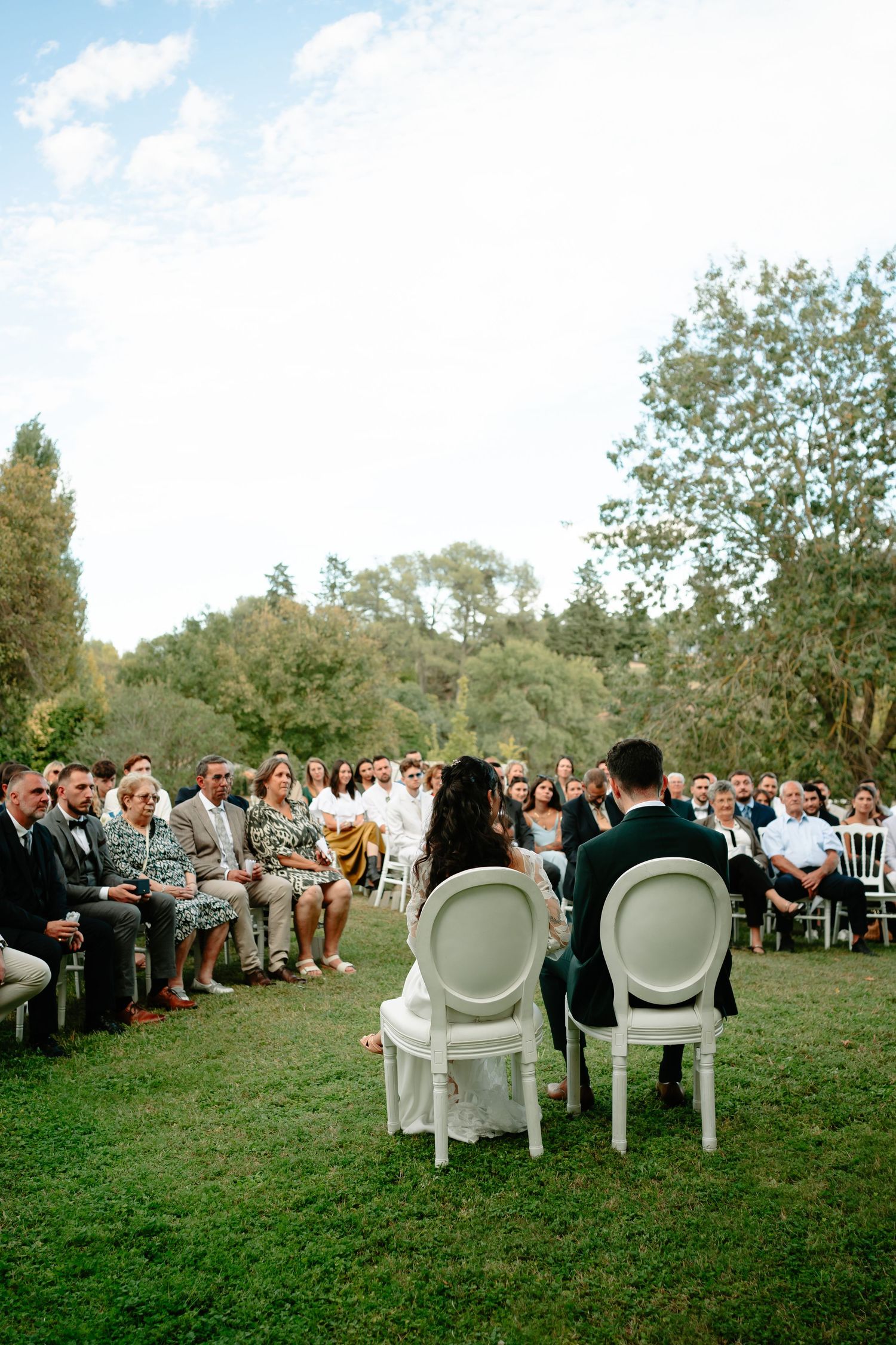 Panoramic view of an outdoor wedding ceremony with white chairs set up on a grassy lawn surrounded by trees.