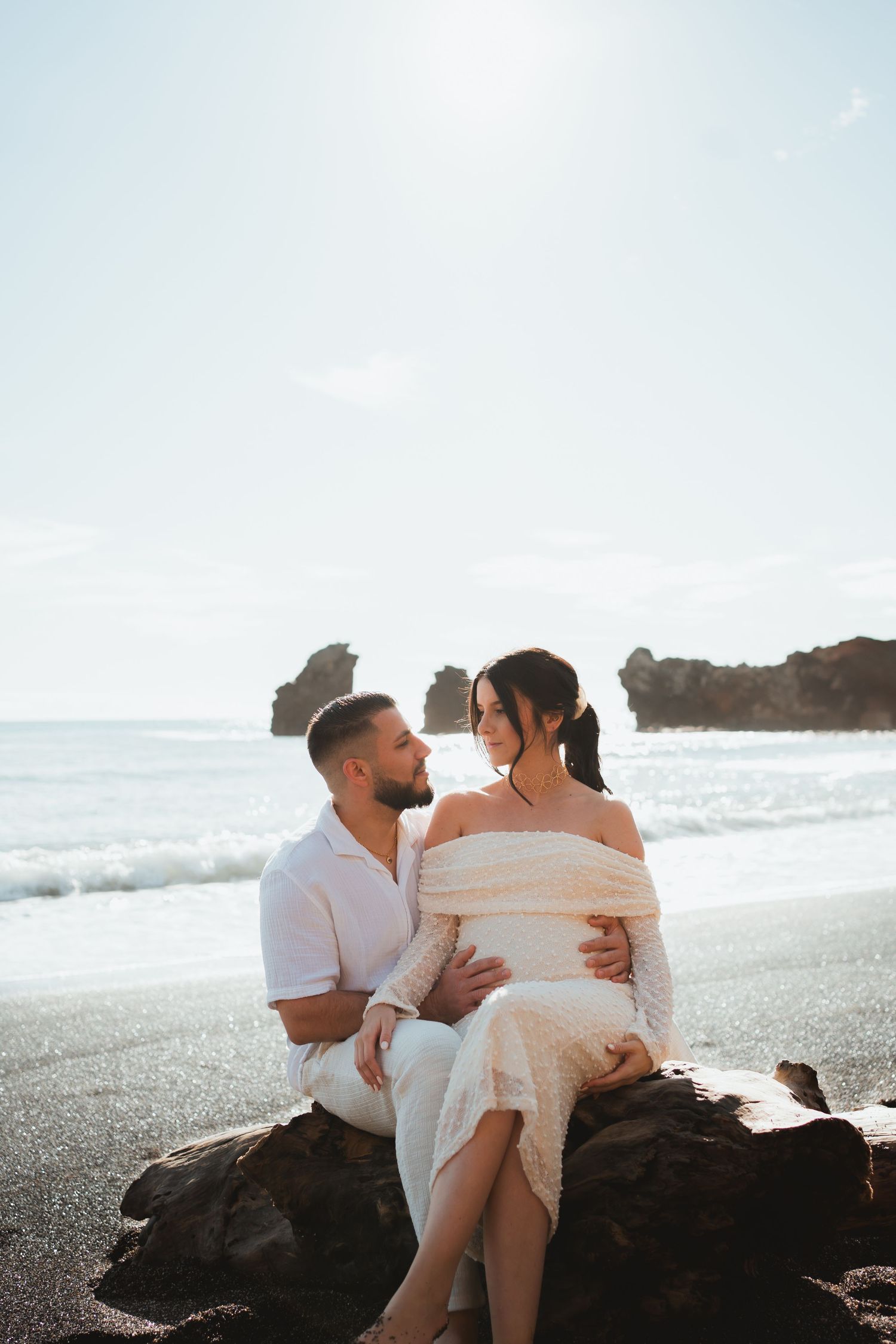 A romantic couple sits on coastal rocks at sunset with ocean waves and cliffs in the background.