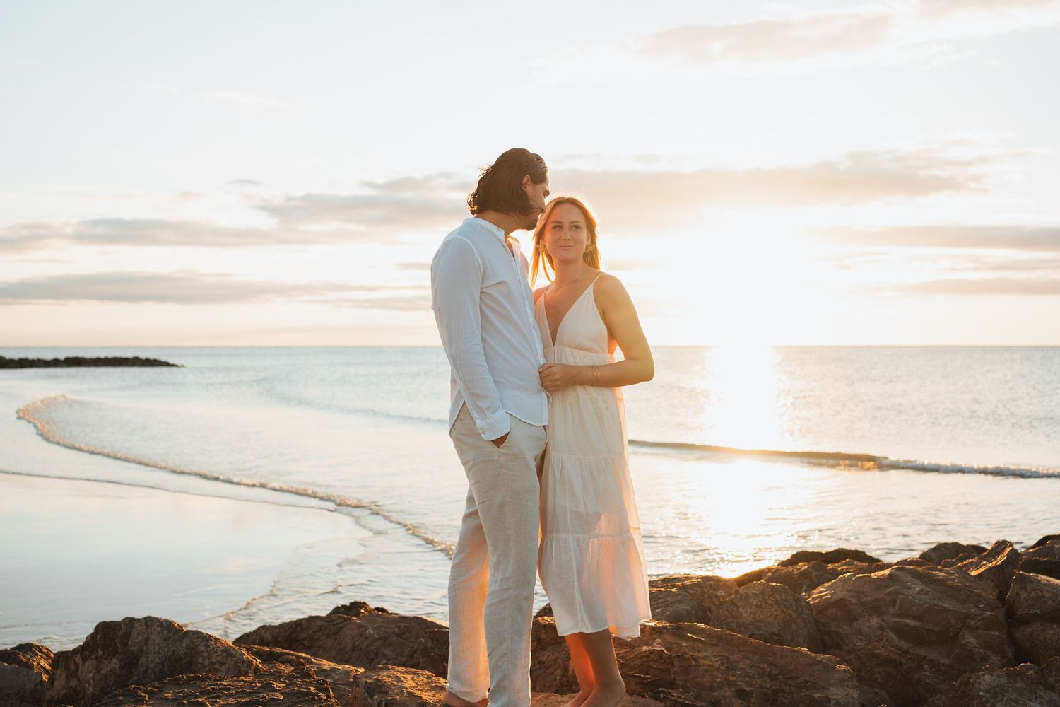 A couple stands together on rocky coastal cliffs at sunset with ocean waves in the background.