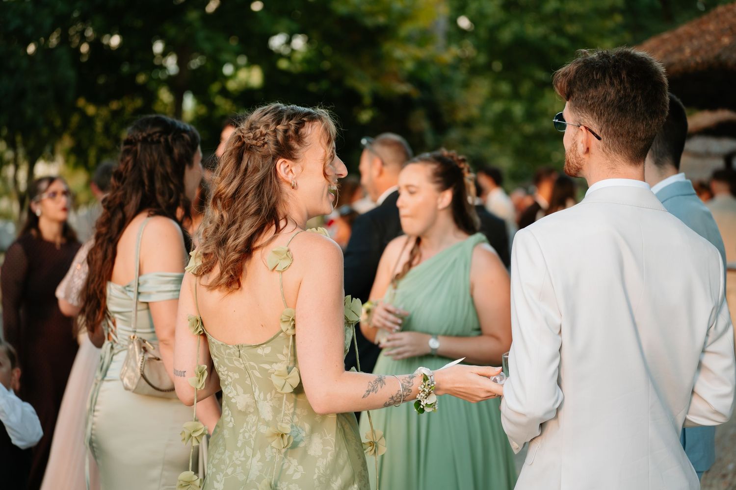 Wedding party members in sage green dresses and suits gather outdoors during a summer celebration.