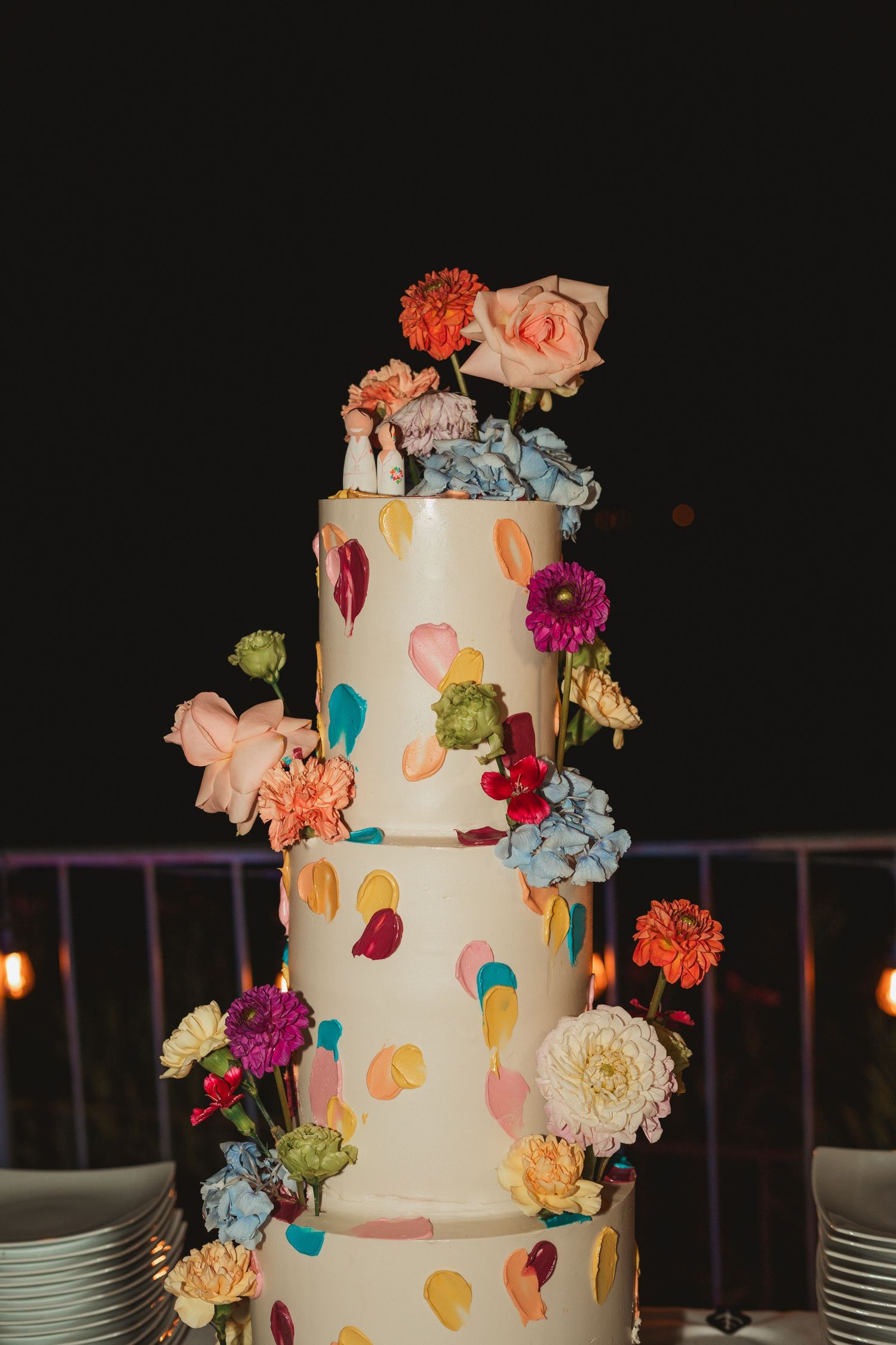 A white wedding cake decorated with colorful fresh flowers and painted brush strokes in pastel colors shown from multiple angles.
