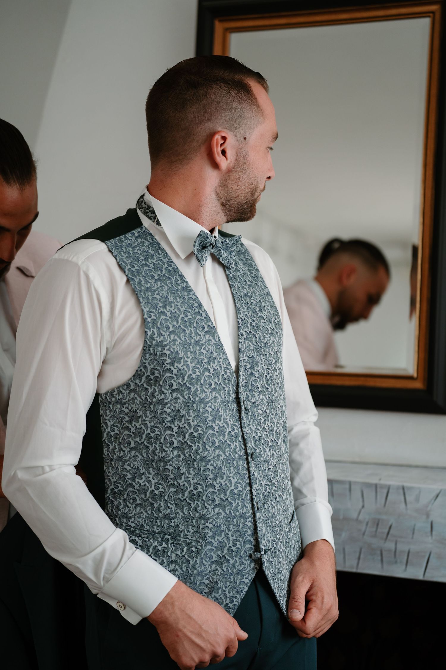 Groom wearing a patterned grey vest and white dress shirt adjusts his bowtie while getting ready in front of a mirror.