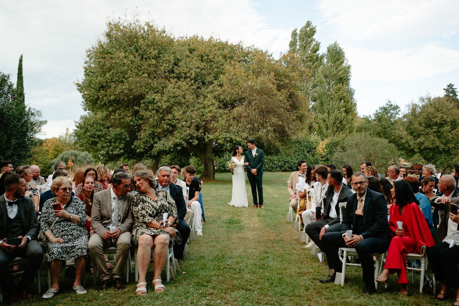 Wedding guests gather outdoors on a grassy hillside for an intimate ceremony surrounded by lush greenery and trees.