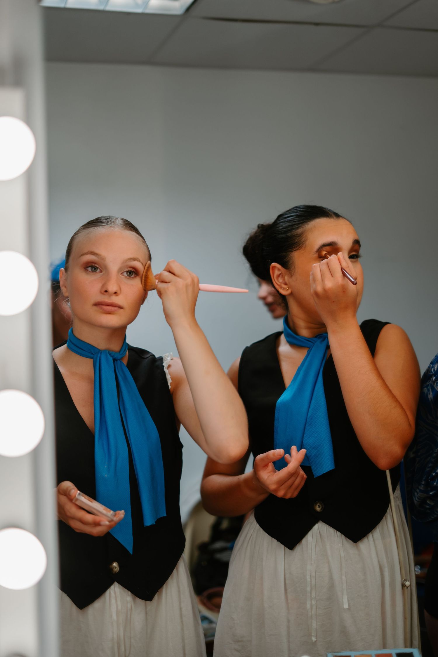 Two performers in matching blue scarves and black vests apply makeup in front of a dressing room mirror.