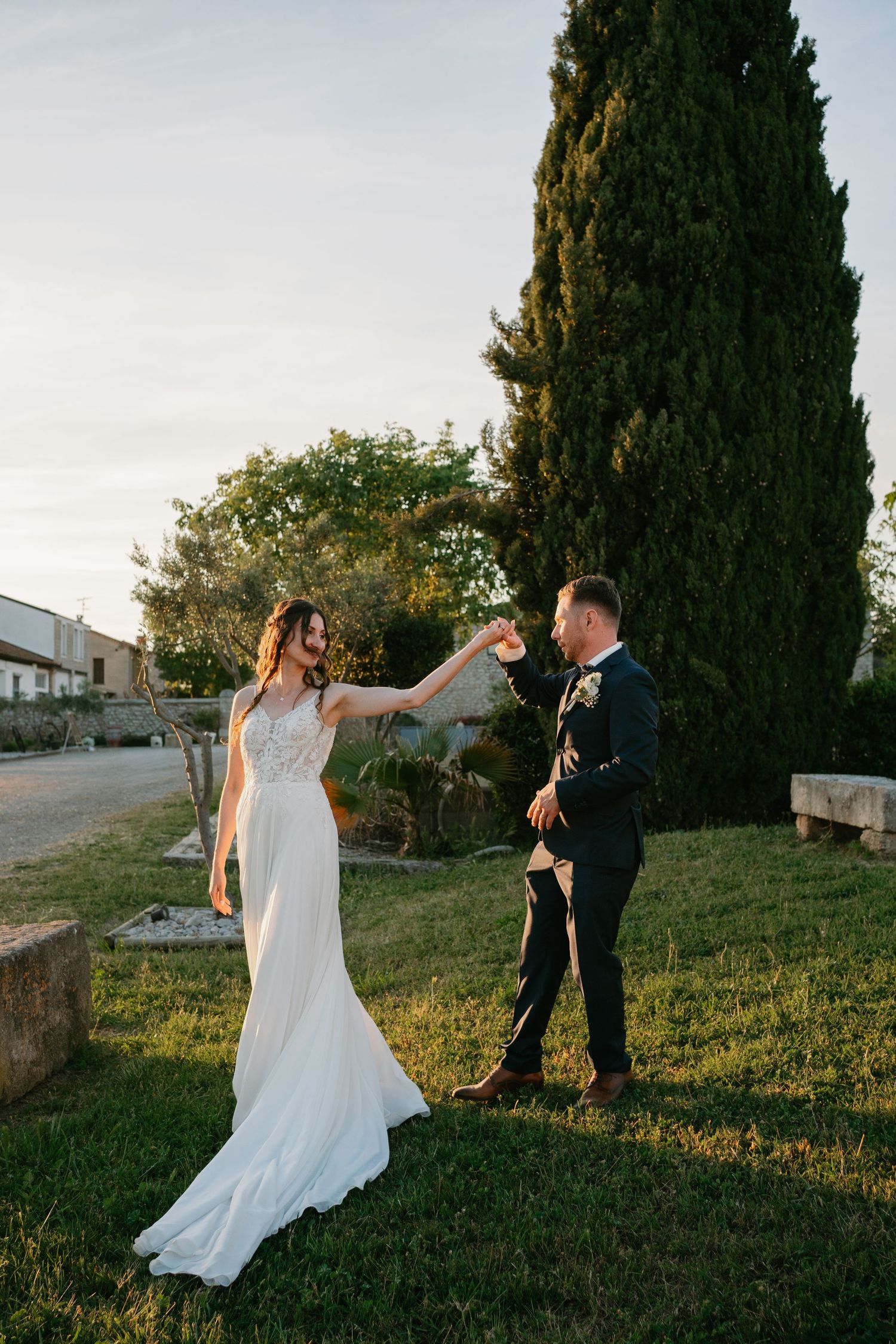 Bride and groom dancing outdoors at golden hour near cypress tree, bride in elegant white gown.