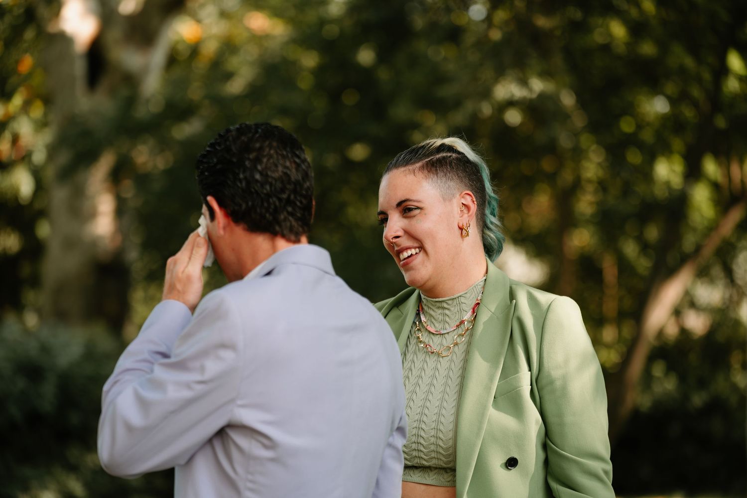 Two people share a candid moment outdoors with one wearing a mint green blazer and knit top in natural lighting.