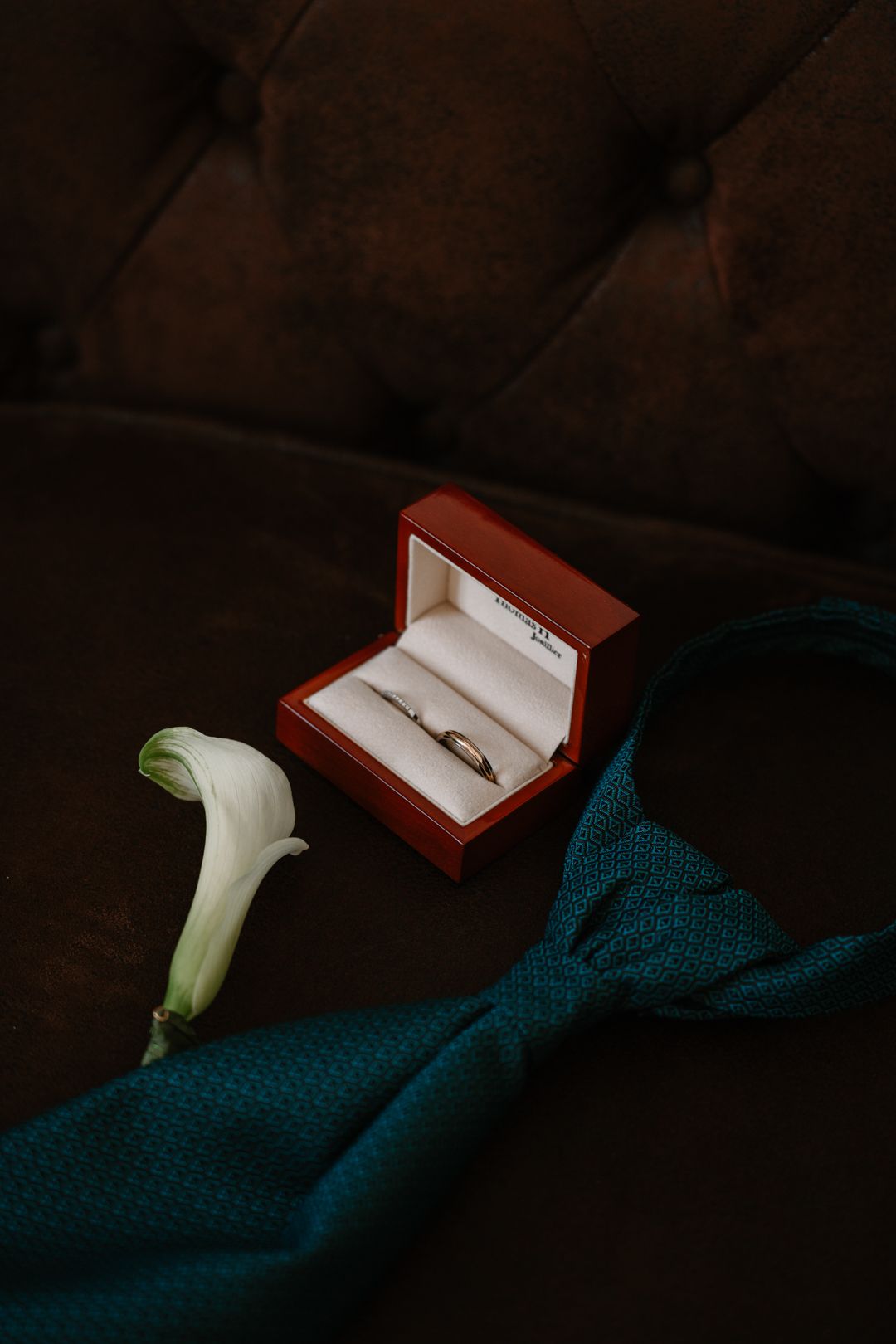 Red jewelry box with wedding rings beside a white calla lily and teal tie on dark brown tufted leather.