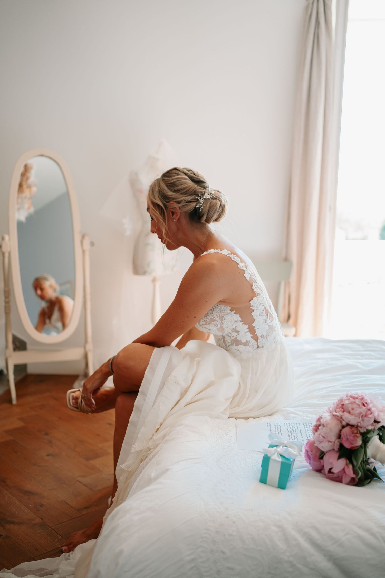 A bride in a white lace wedding dress sits on a bed with pink flowers and turquoise decor in a bright room.