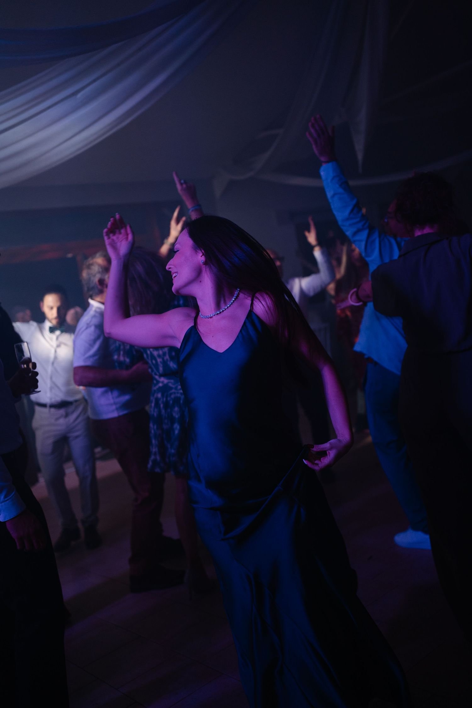 People dancing and celebrating at a dimly lit party with blue and purple lighting effects in a tent venue.