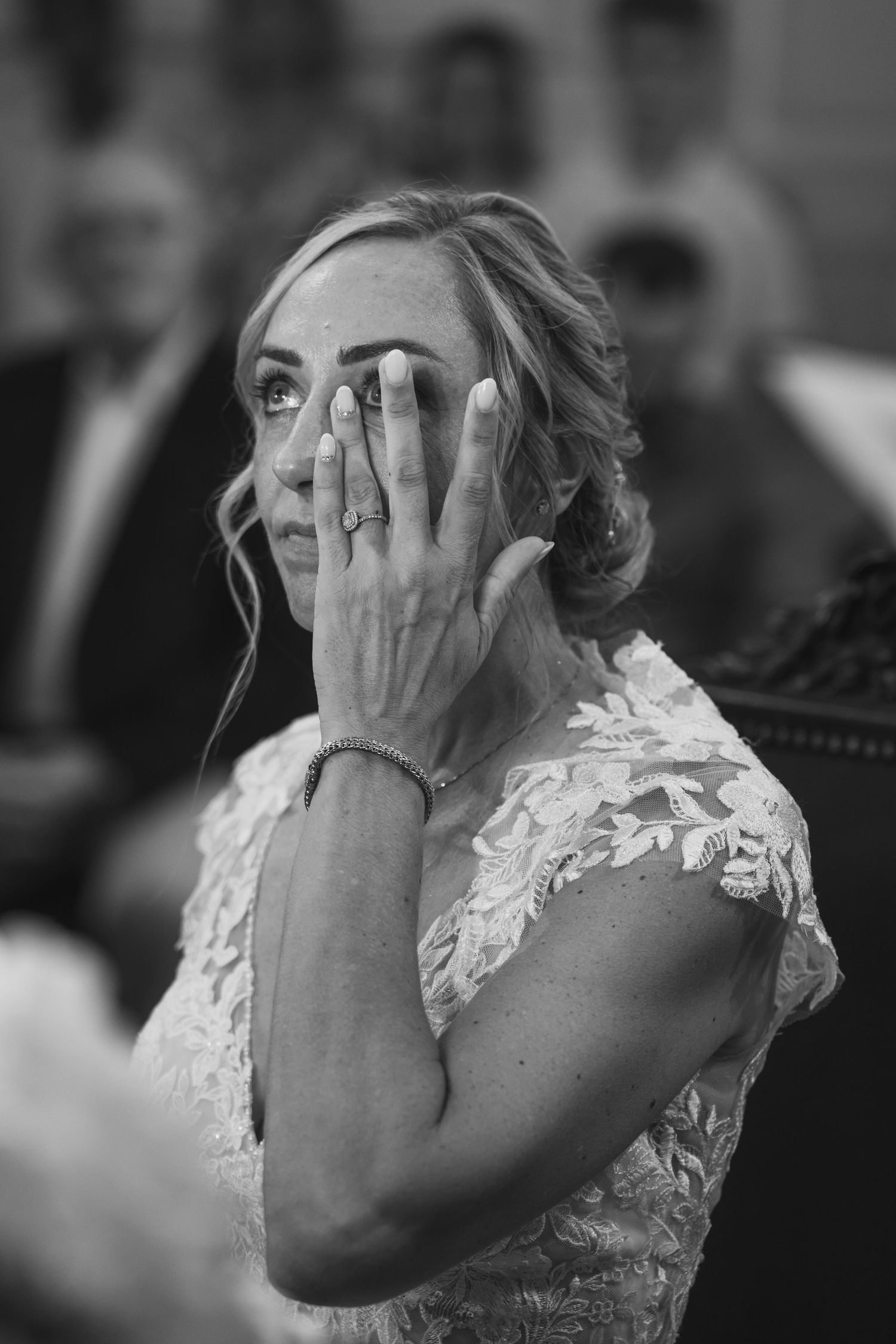 A bride in a white lace dress covers her face with her hand in a black and white emotional wedding moment.