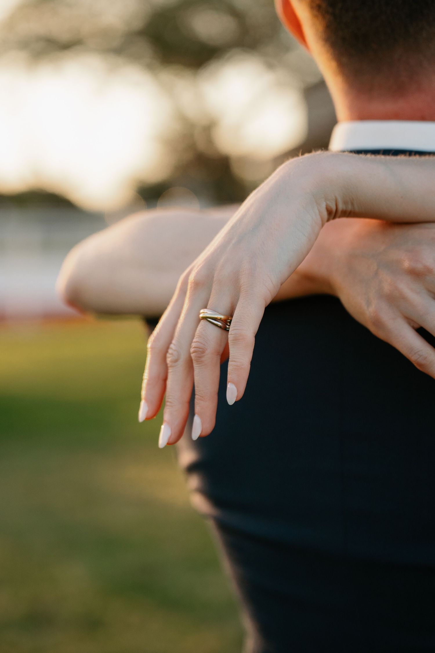Close-up of a woman's hand with a gold ring resting on a man's shoulder in a romantic outdoor setting.