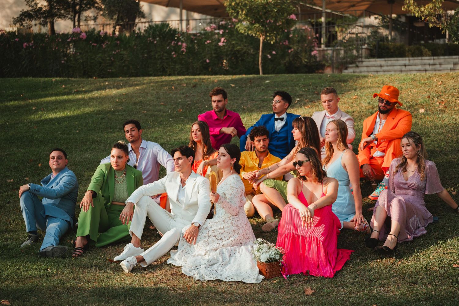 A wedding party relaxes on a grassy lawn during golden hour, wearing colorful attire and enjoying the outdoor setting.
