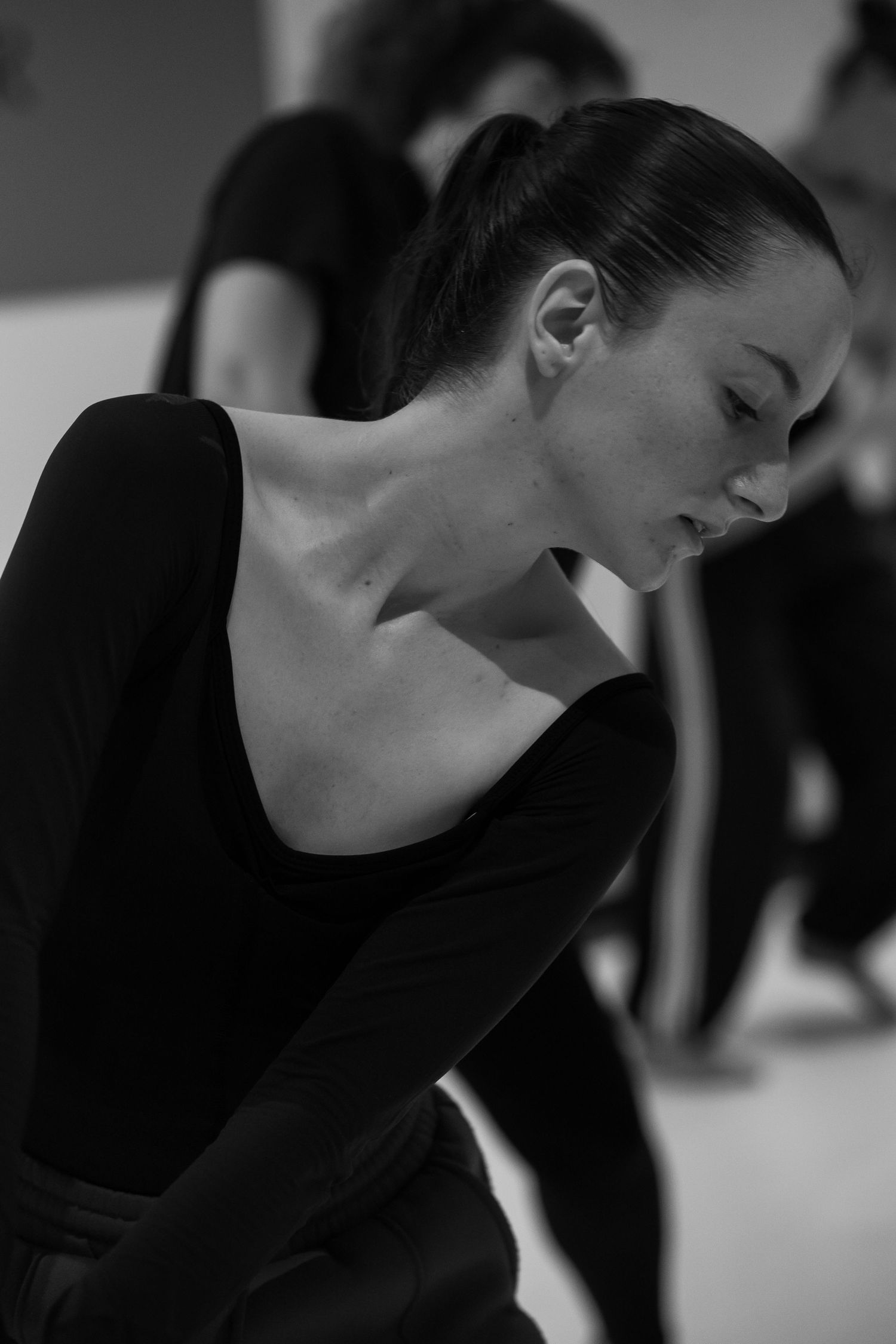 A dancer in a black leotard bends gracefully during practice, captured in an artistic black and white photograph.