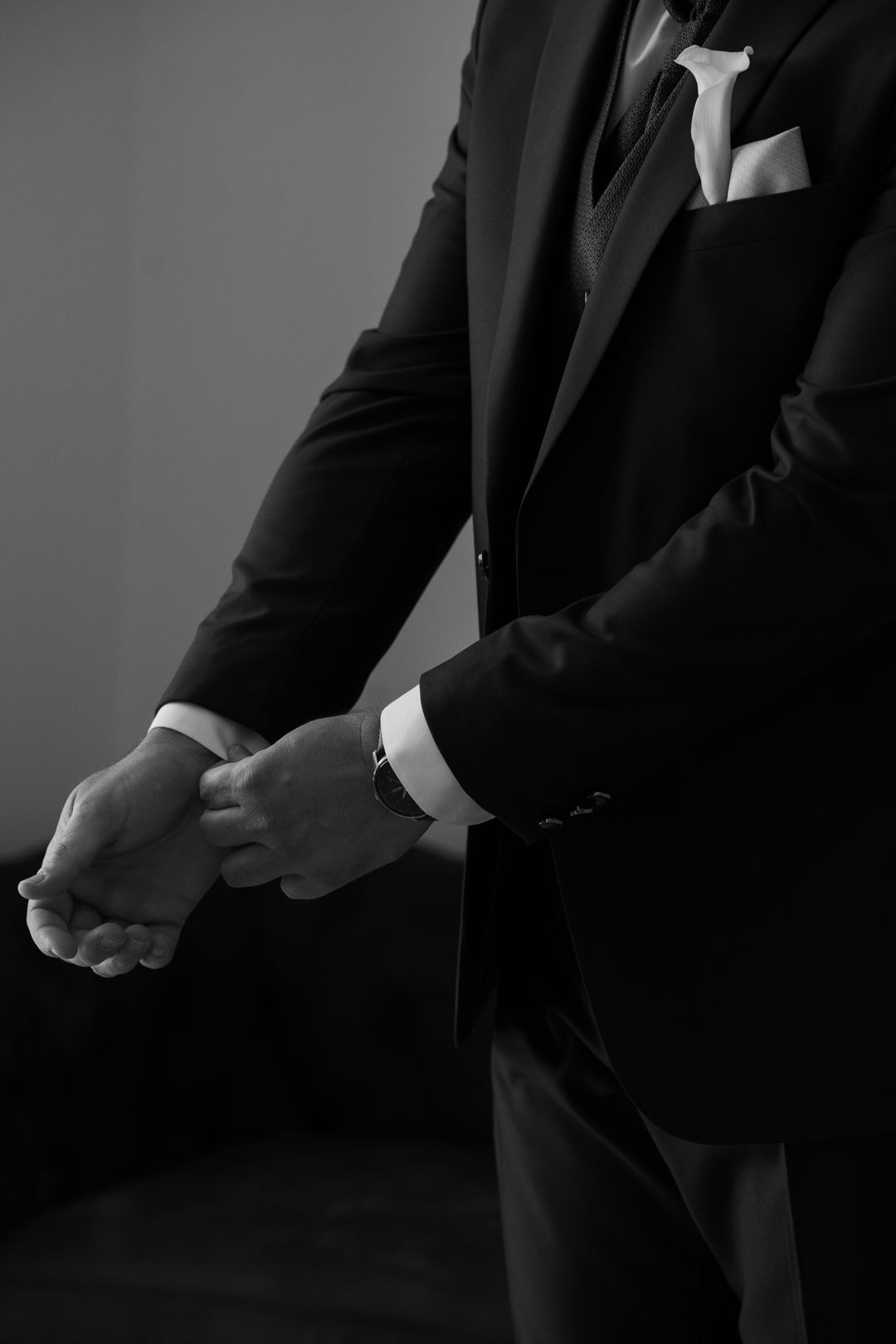 Black and white photo of a groom in a dark suit adjusting his cufflinks, with a white tie and pocket square.
