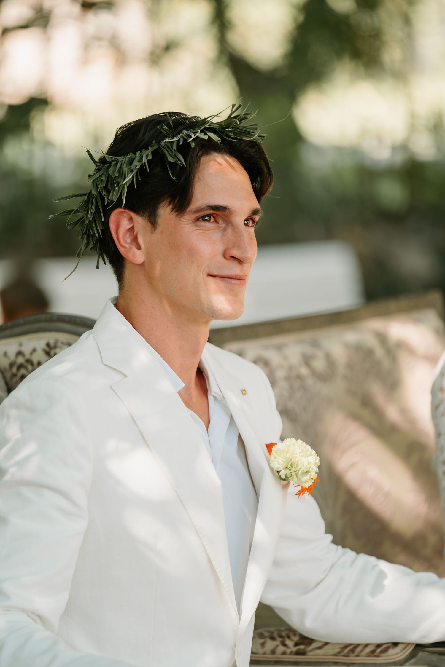 A person in a crisp white suit jacket and laurel crown smiles at a wedding celebration.