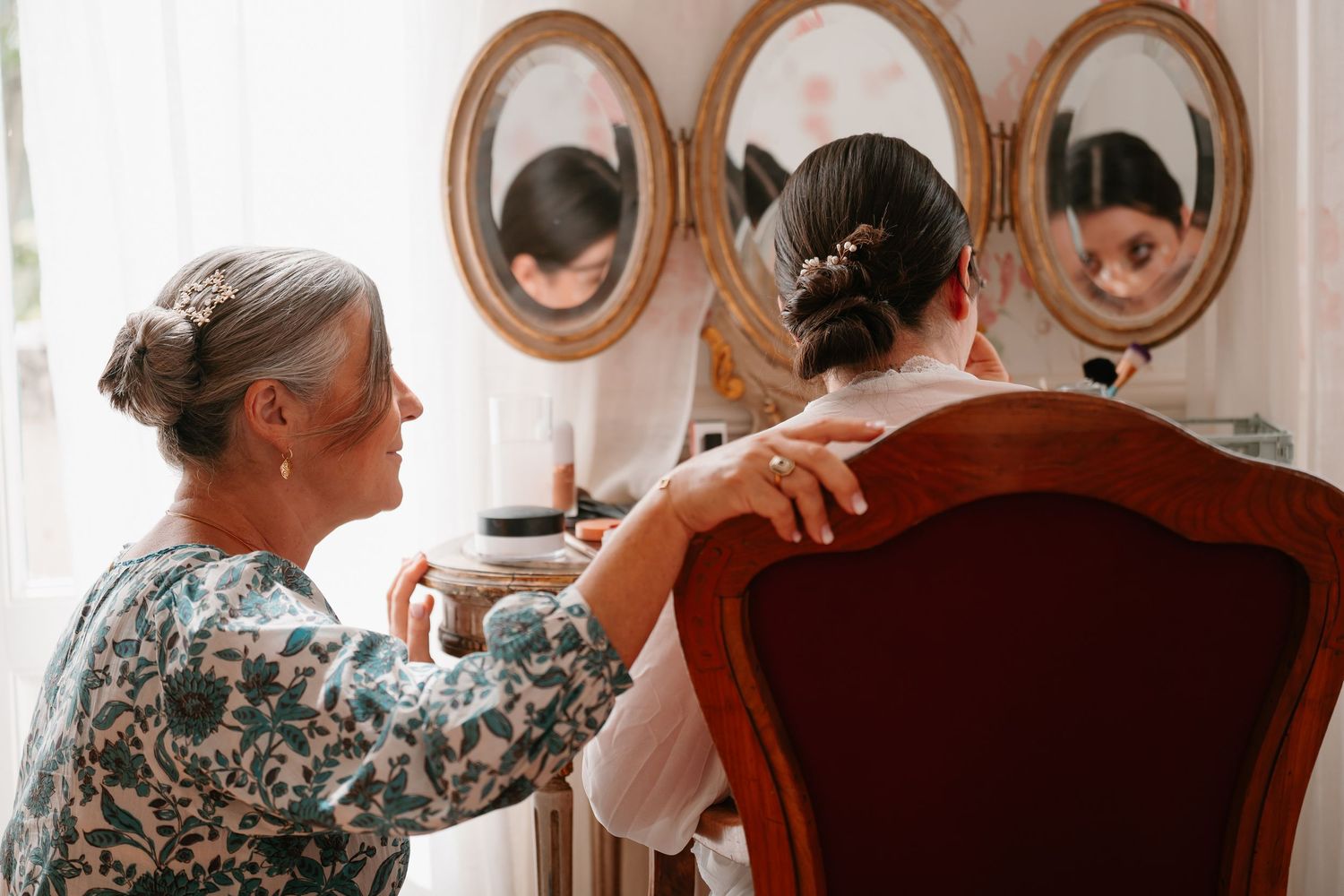 Two people sitting at a wooden dining table share a warm conversation in a sunlit room with oval mirrors on the wall.