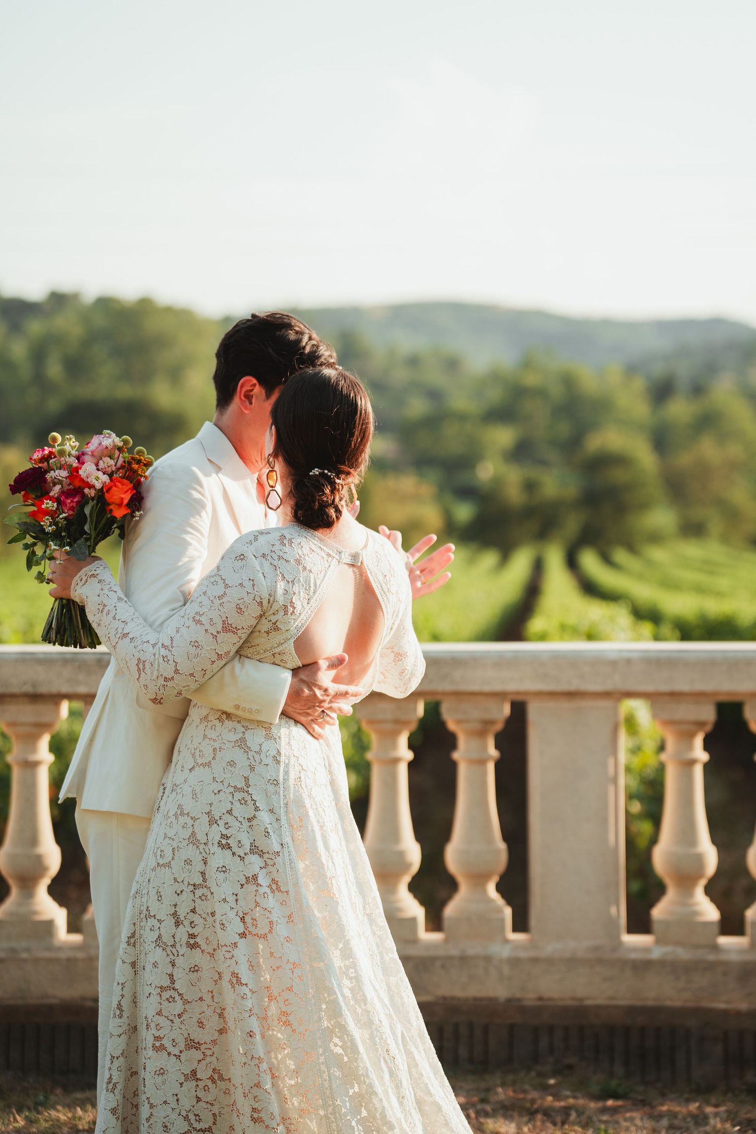 A couple in wedding attire embrace on a balcony overlooking rolling green hills during a golden sunset.
