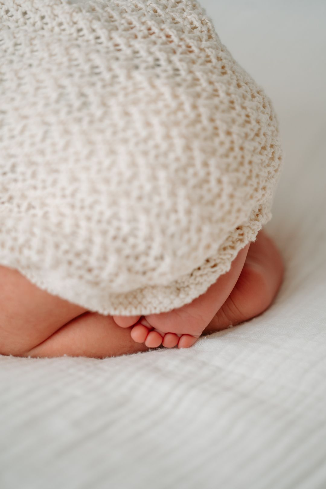 Tiny baby feet peek out from under a delicate white lace blanket on soft white bedding.