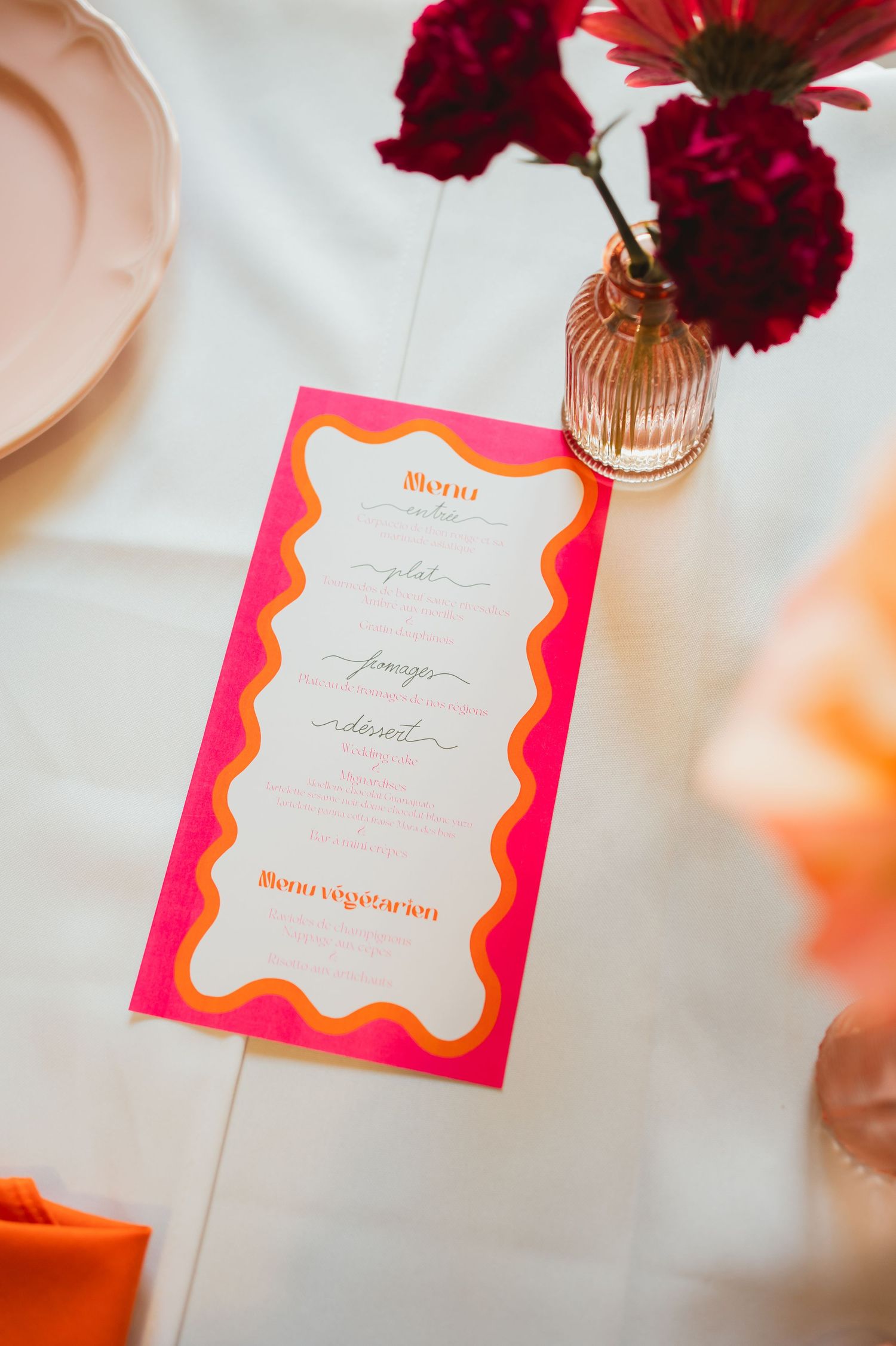Deep red carnations in a gold vase next to elegant pink and gold wedding menu cards on a white tablecloth.