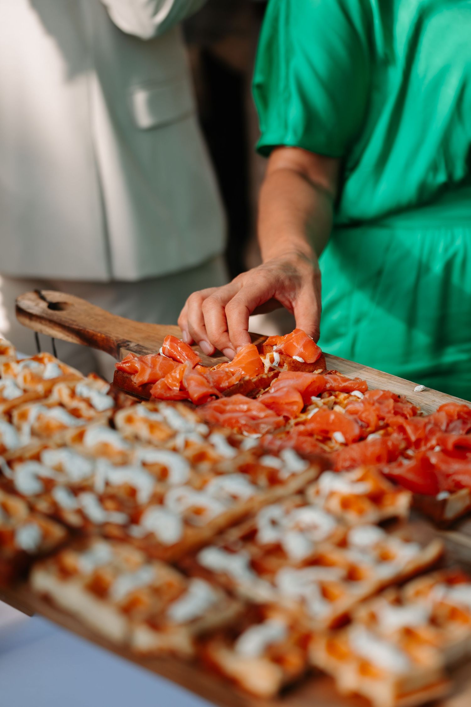 Someone in green prepares appetizers on a wooden cutting board, arranging red toppings in a precise pattern.