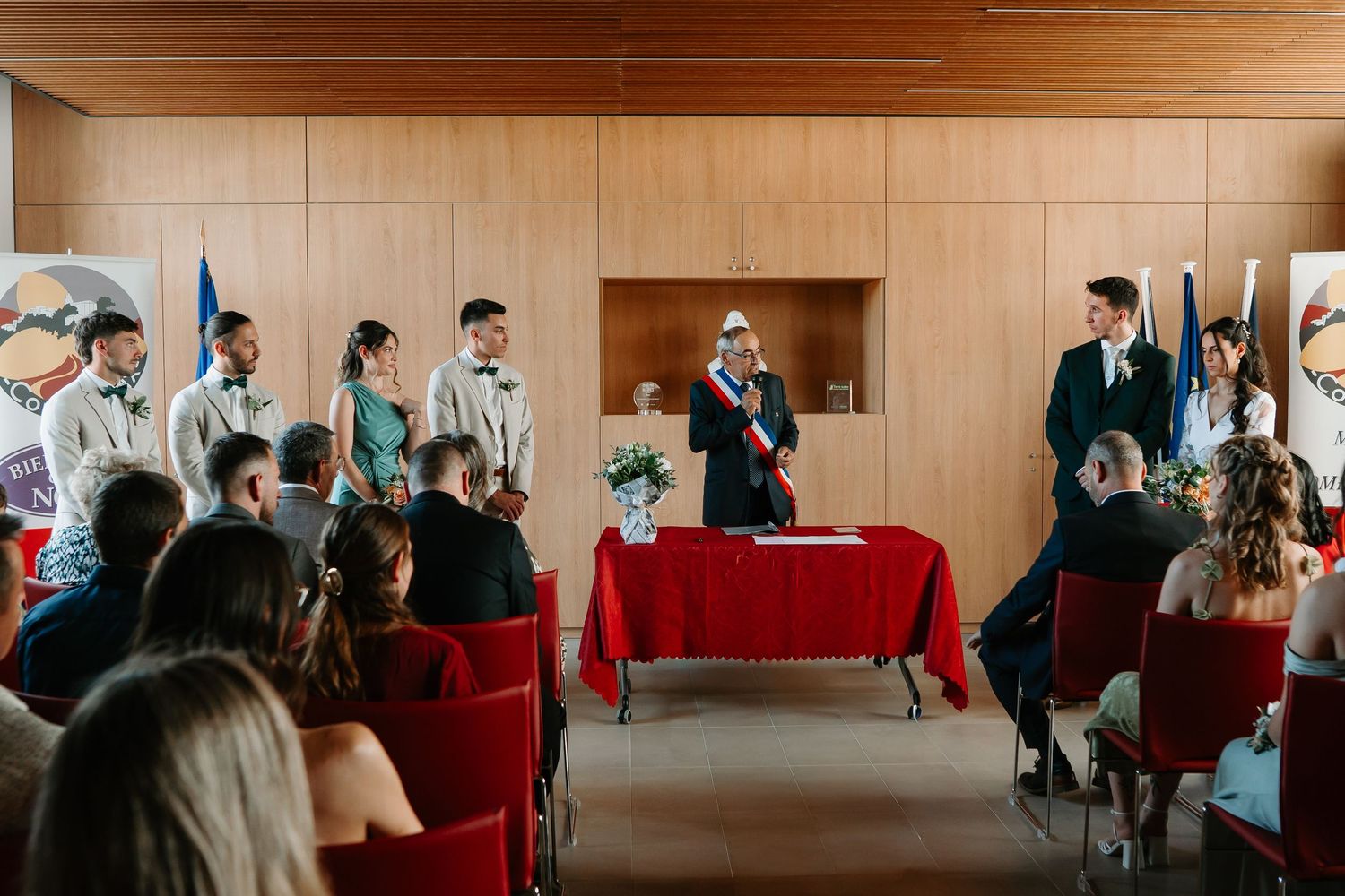 People gather in a large room with wooden paneled walls and red chairs arranged in rows for an event.