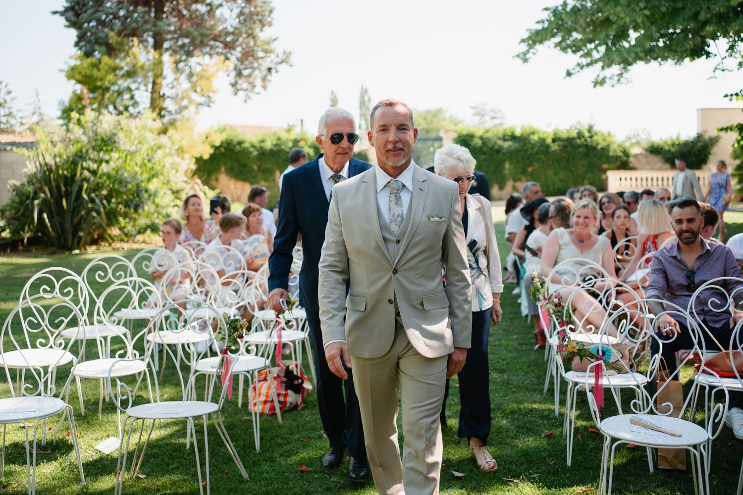 Wedding ceremony setup outdoors with white metal chairs on a sunny lawn with guests and attendees present.
